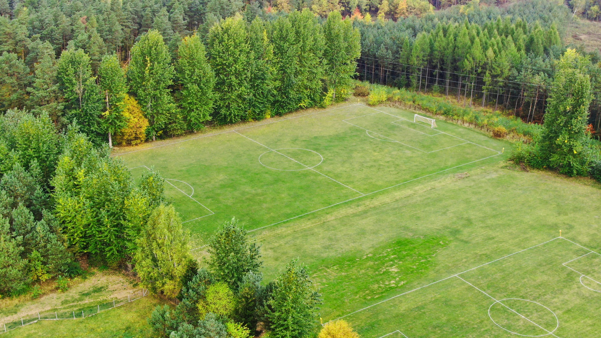 Widok z lotu ptaka na zielone boisko do piłki nożnej otoczone lasem, z widocznymi bramkami i liniami wyznaczającymi pole gry.