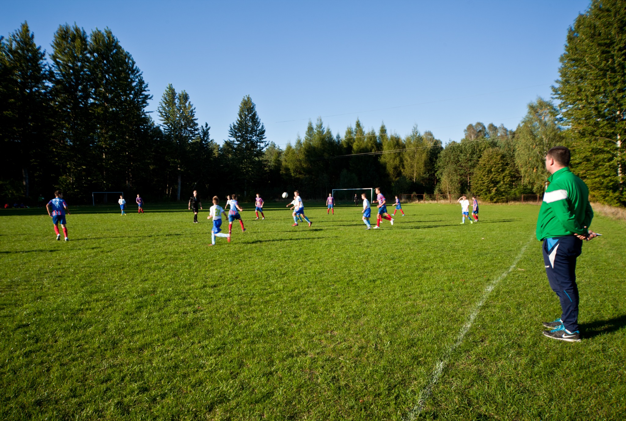 Mecz piłki nożnej na trawiastym boisku, zawodnicy w ruchu, trener w zielonej kurtce obserwuje z boku