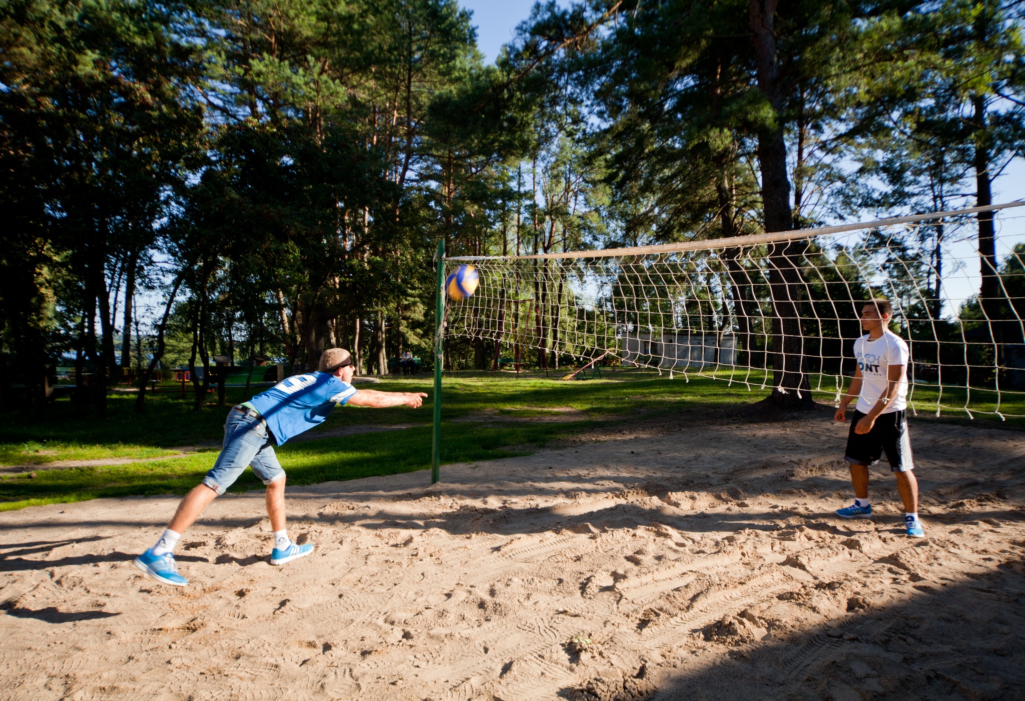Dwóch mężczyzn gra w siatkówkę na piaszczystym boisku otoczonym drzewami; jeden w niebieskiej koszulce w trakcie uderzenia piłki, drugi w białej koszulce obserwuje.