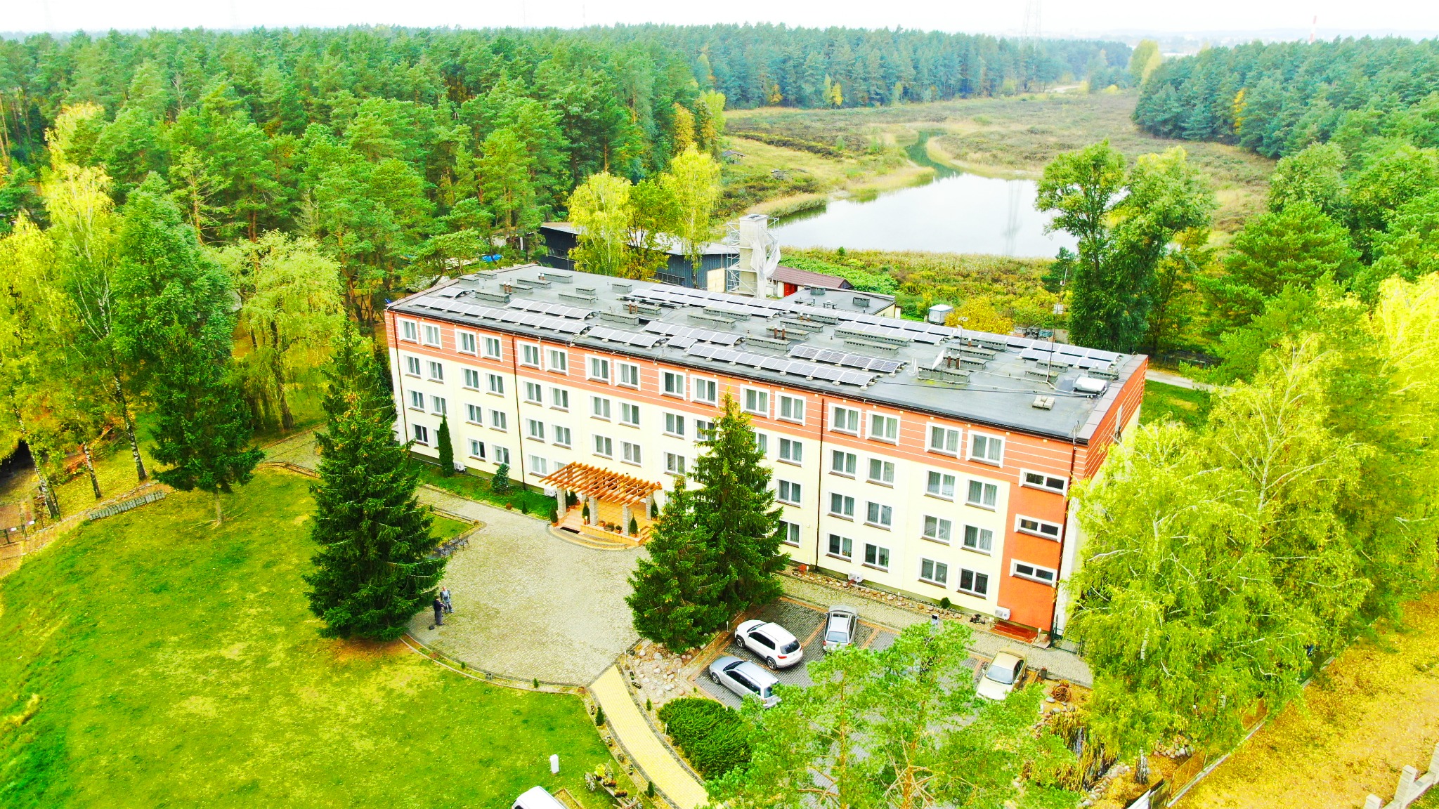 Widok z lotu ptaka na trzypiętrowy pensjonat z panelami słonecznymi na dachu, otoczony zielonymi drzewami i niewielkim jeziorem w tle.