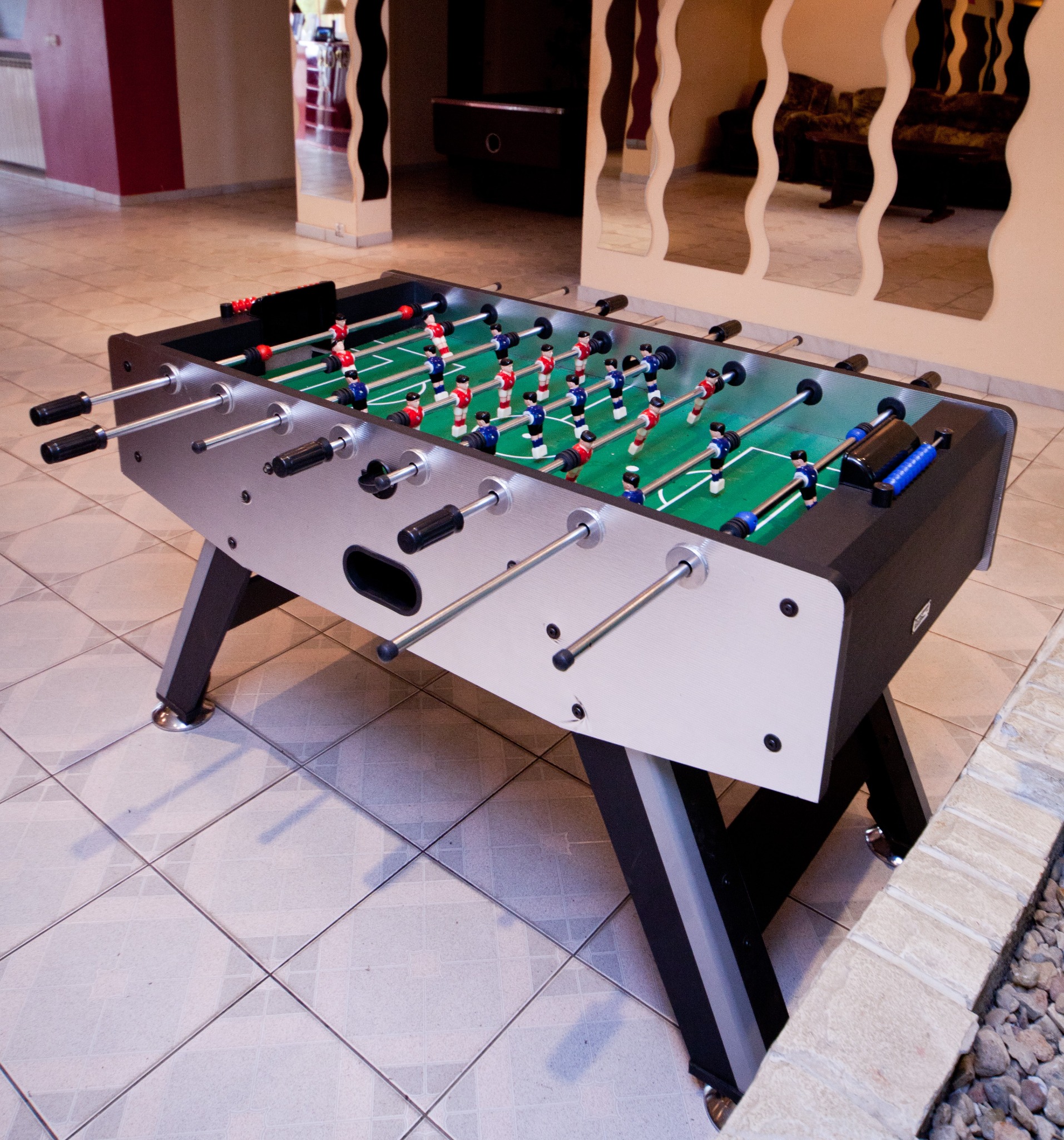 Stół do piłkarzyków z figurkami w kolorach czerwonym i niebieskim, ustawiony w pomieszczeniu z beżowymi płytkami i dekoracyjnymi lustrami na ścianie.