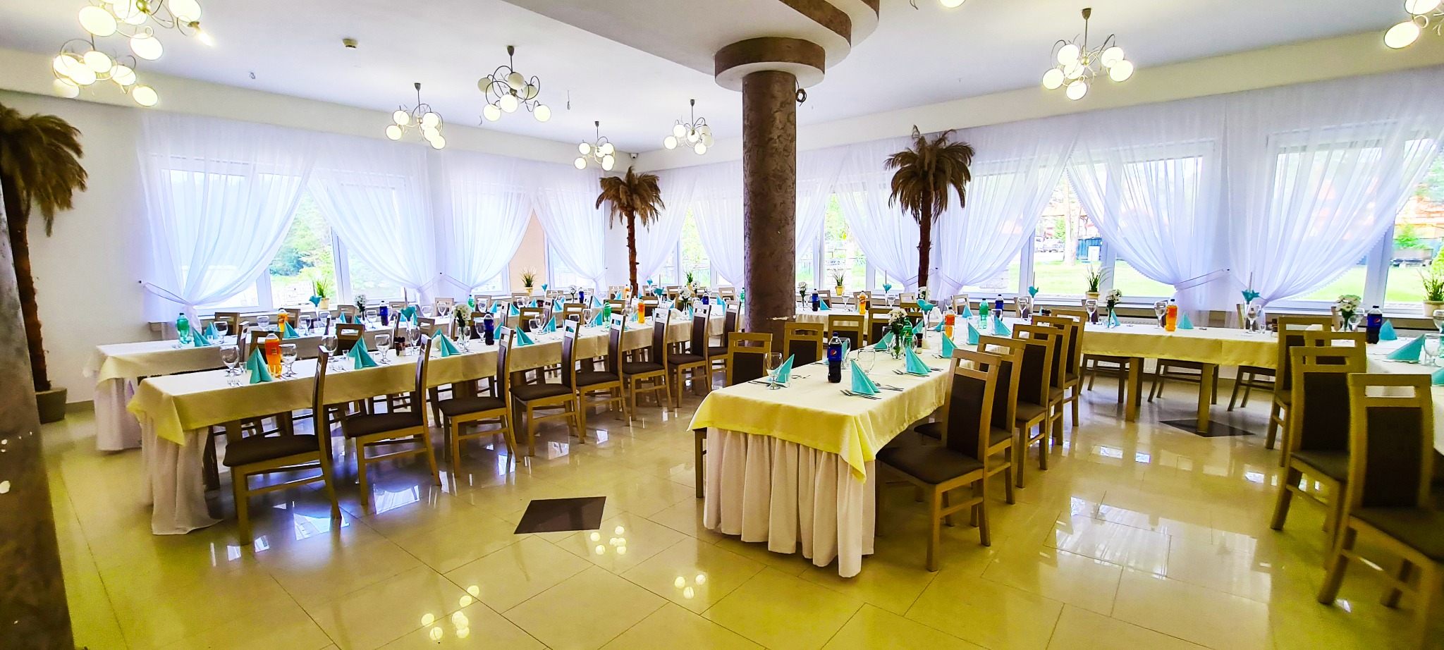 Sala bankietowa z nakrytymi stołami, krzesłami, dekoracjami z palmami w donicach i białymi firanami w oknach, gotowa na przyjęcie gości.