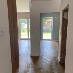 Mariusz PGAM - Ułożona parkietowa podłoga w jodełkę w jasnym wnętrzu z oknami na ogród. Widoczny fragment ściany z surowej cegły i przygotowane otwory drzwiowe.