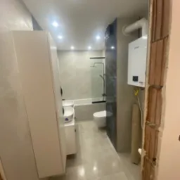 Wykończona łazienka z białymi meblami, grzejnikiem Vaillant i kabiną prysznicową, sfotografowana z perspektywy surowego otworu drzwiowego.