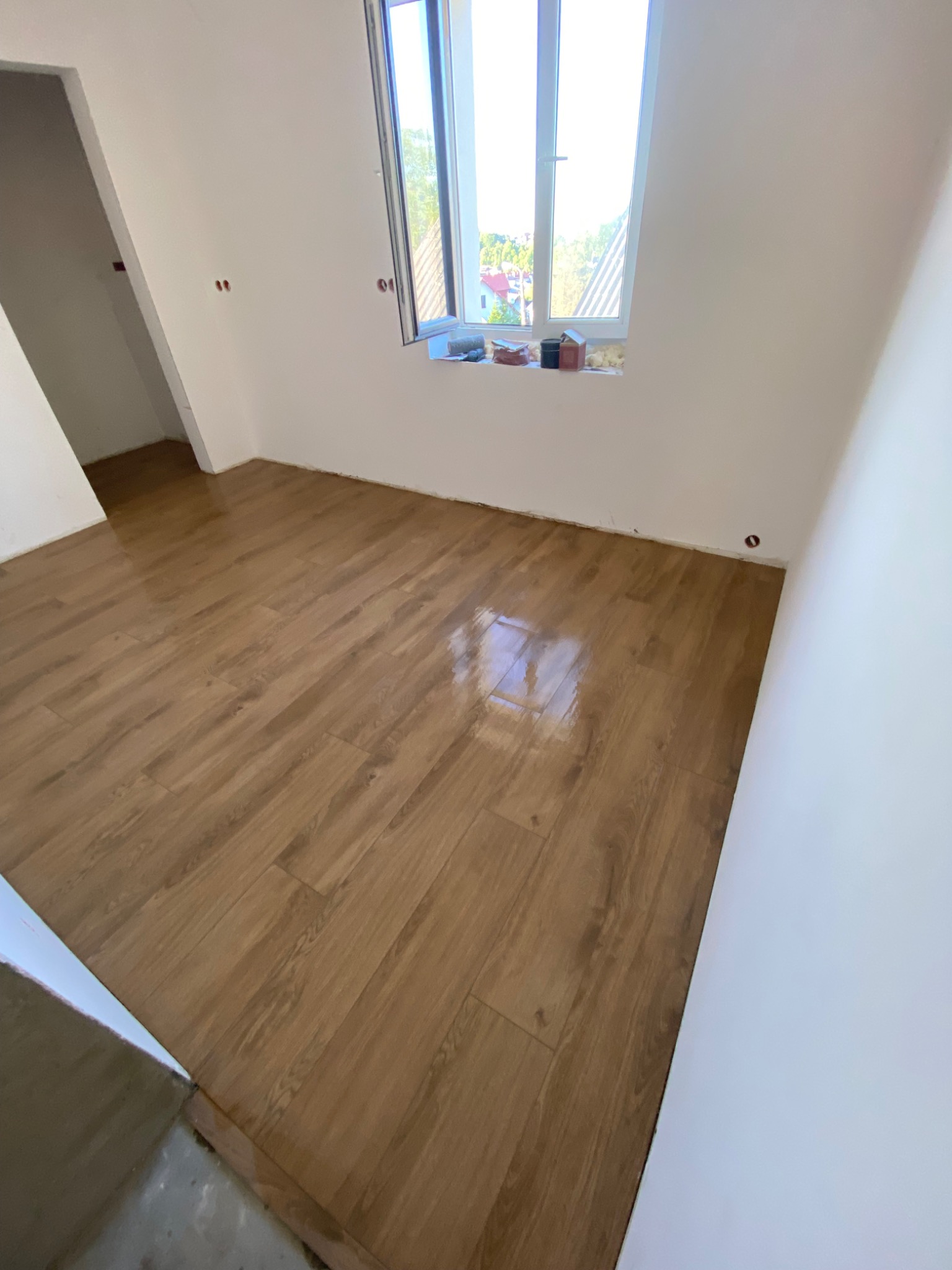 Jasne panele podłogowe ułożone w pokoju z białymi ścianami i oknem. Widoczne detale wykończenia i świeżo pomalowane ściany. Kąt ujęcia podkreśla przestronność.