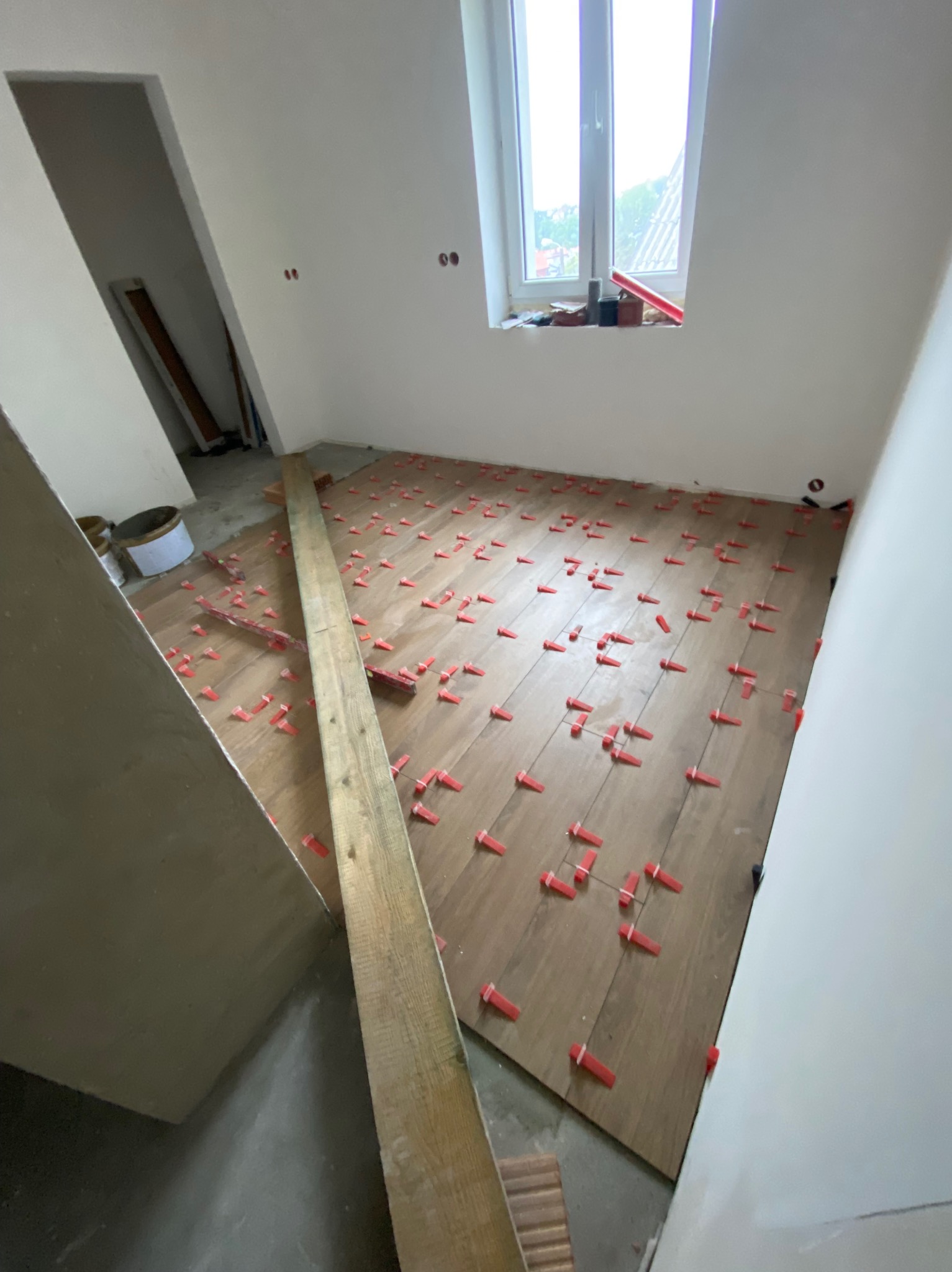 Układanie podłogi z paneli drewnopodobnych z użyciem czerwonych klinów dystansowych w jasnym pomieszczeniu z oknem. Widoczna deska i przygotowane podłoże.