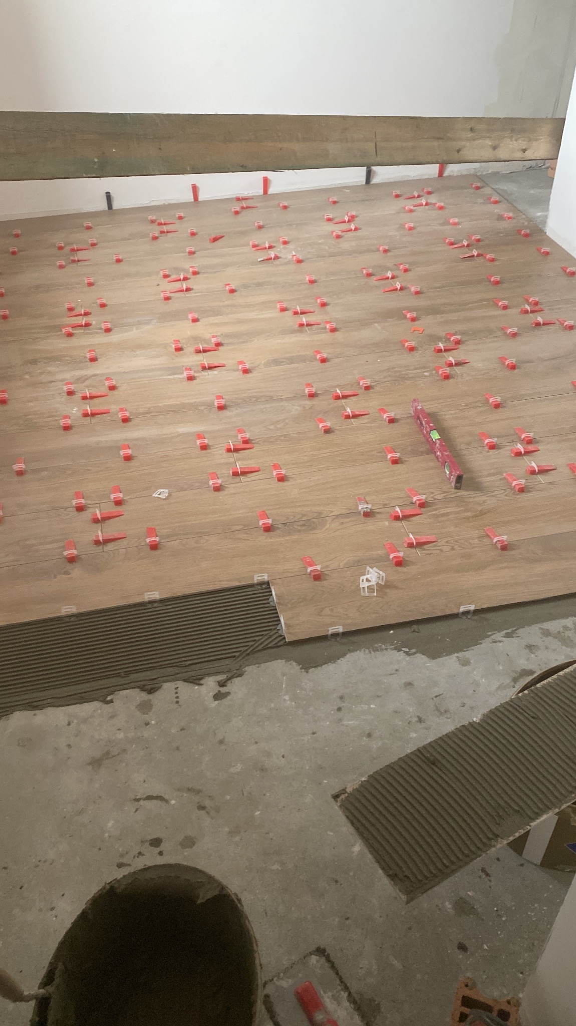 Układanie płytek podłogowych imitujących drewno. Widoczne czerwone kliny poziomujące i poziomica. Częściowo położone płytki na kleju. Wiadro z zaprawą w rogu.
