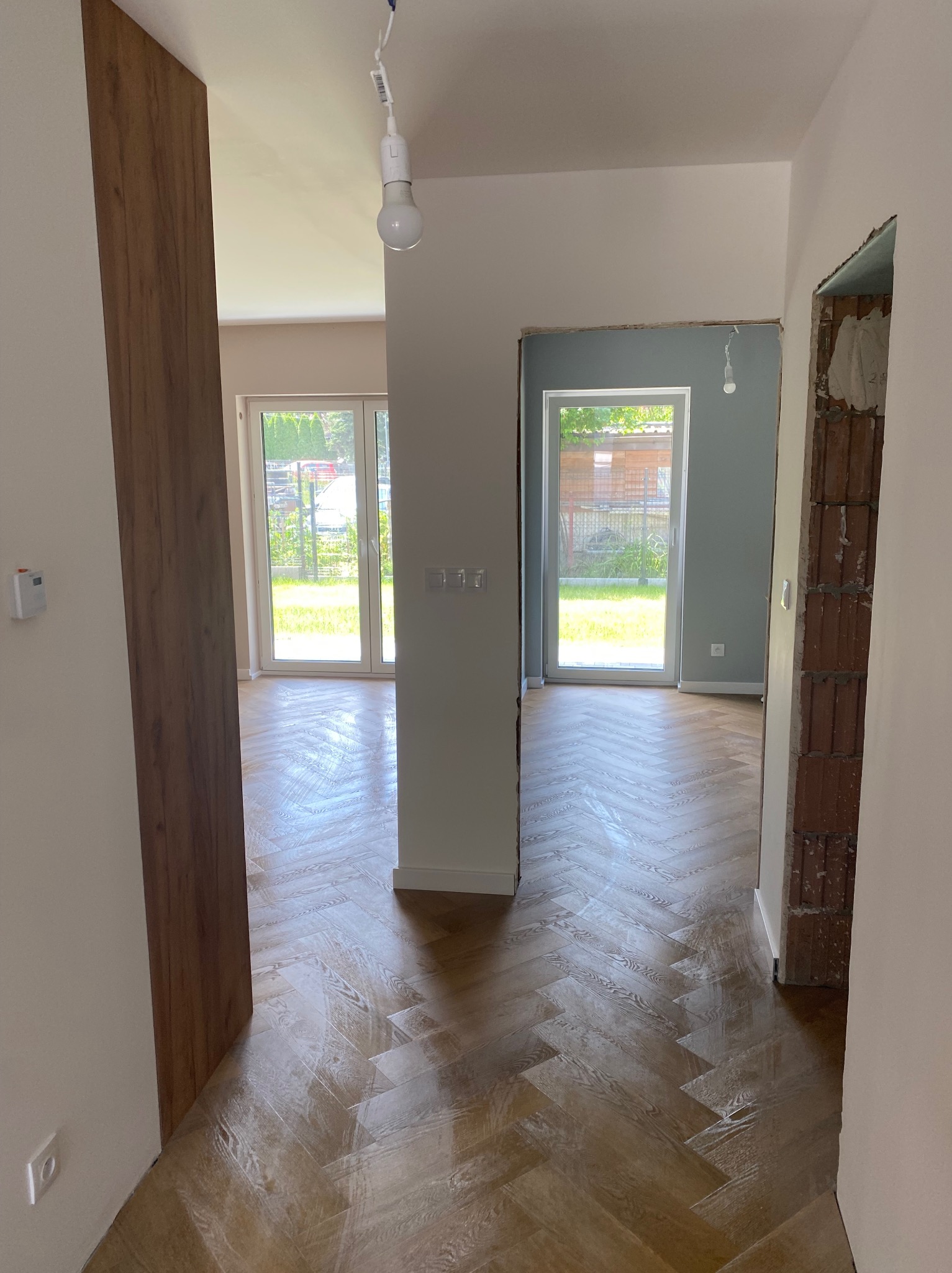 Ułożona parkietowa podłoga w jodełkę w jasnym wnętrzu z oknami na ogród. Widoczny fragment ściany z surowej cegły i przygotowane otwory drzwiowe.