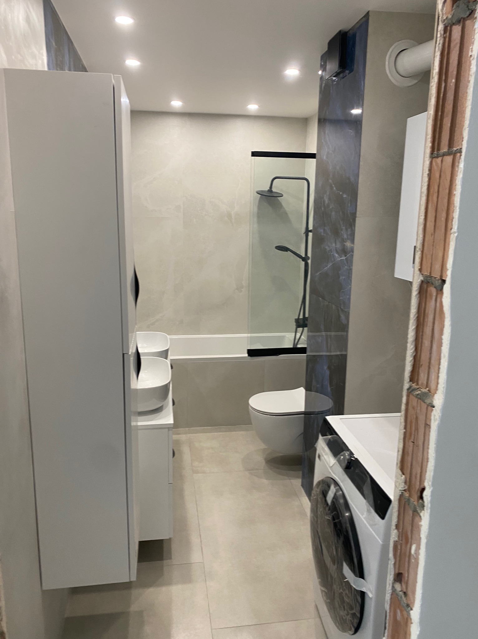 Nowoczesna łazienka z kabiną prysznicową, umywalką na szafce, toaletą i pralką. Widoczne płytki w odcieniach szarości i bieli, oraz wykończona ściana z cegły.