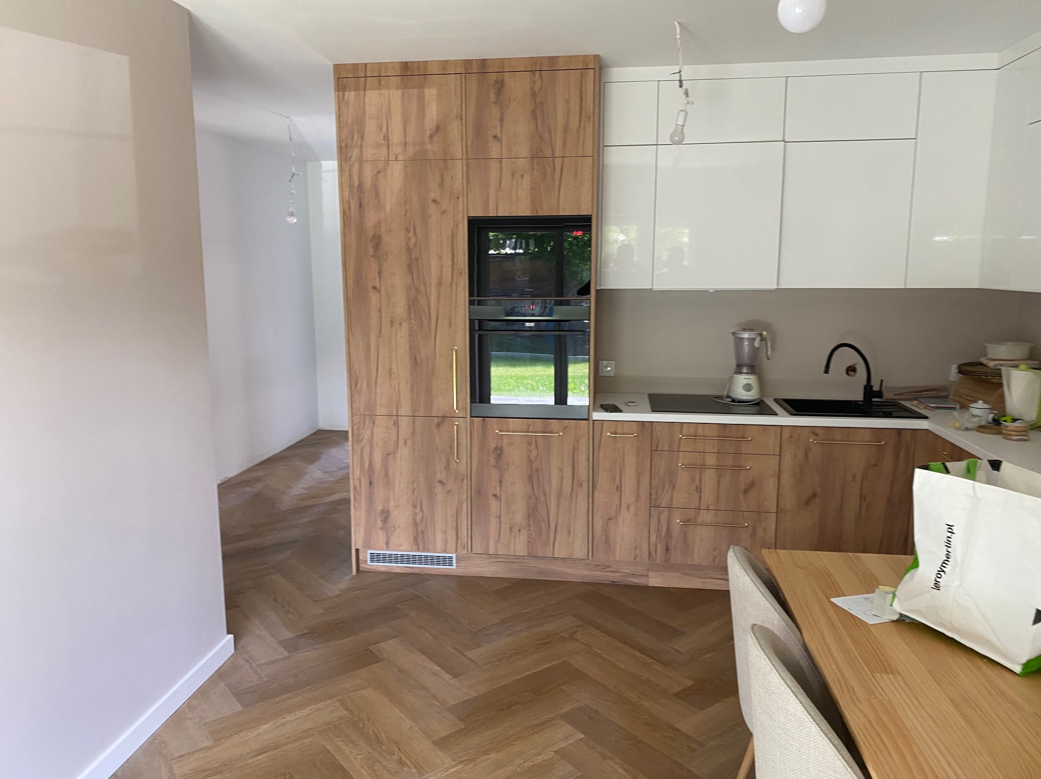 Nowoczesna kuchnia z drewnianymi frontami szafek, białymi górnymi szafkami i parkietem w jodełkę. Widoczny sprzęt AGD i fragment stołu z krzesłami.