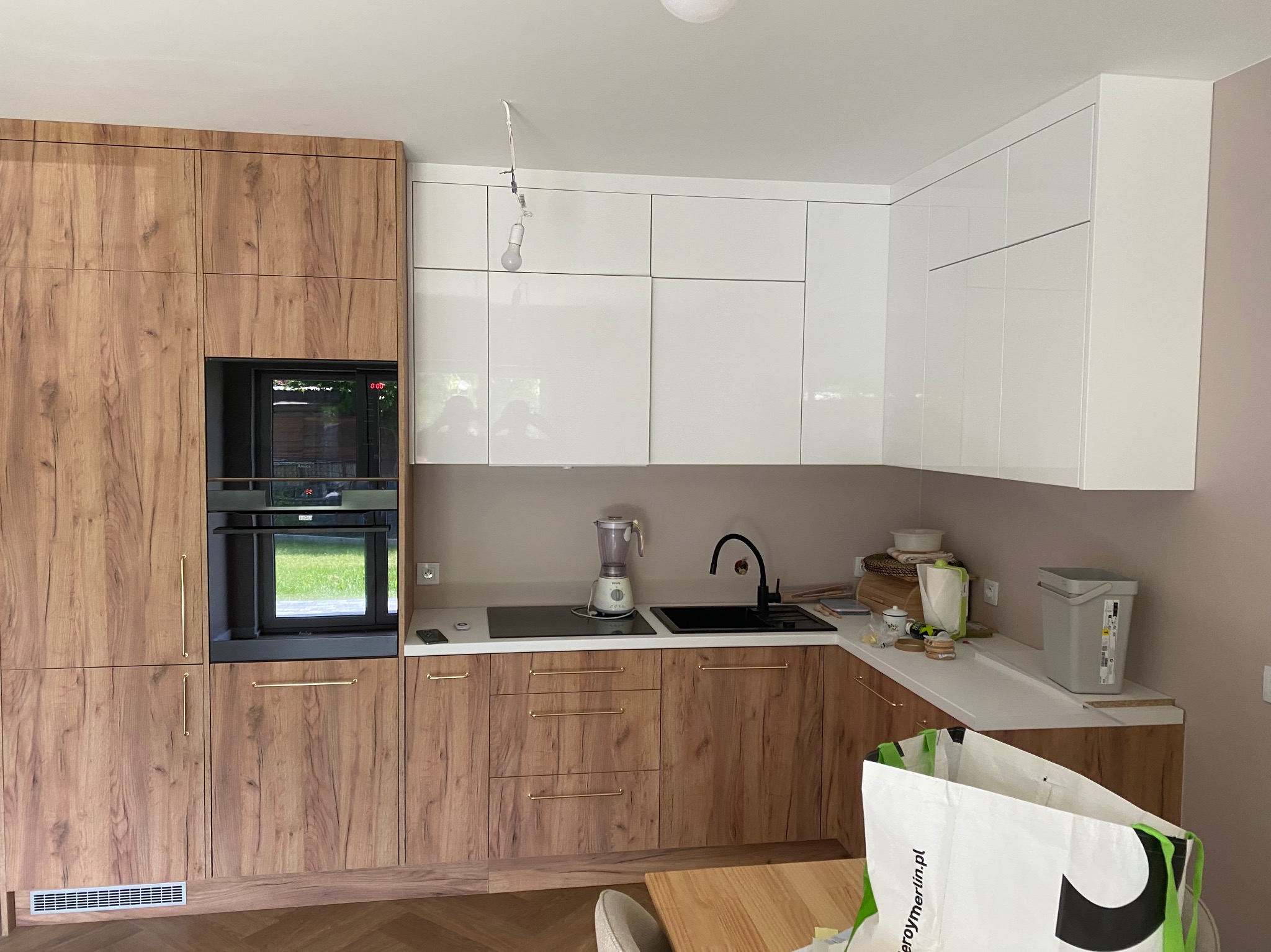 Ukończona kuchnia z drewnianymi szafkami i białymi frontami górnymi. Widoczny sprzęt AGD i armatura. Na blacie kuchennym stoją akcesoria kuchenne.