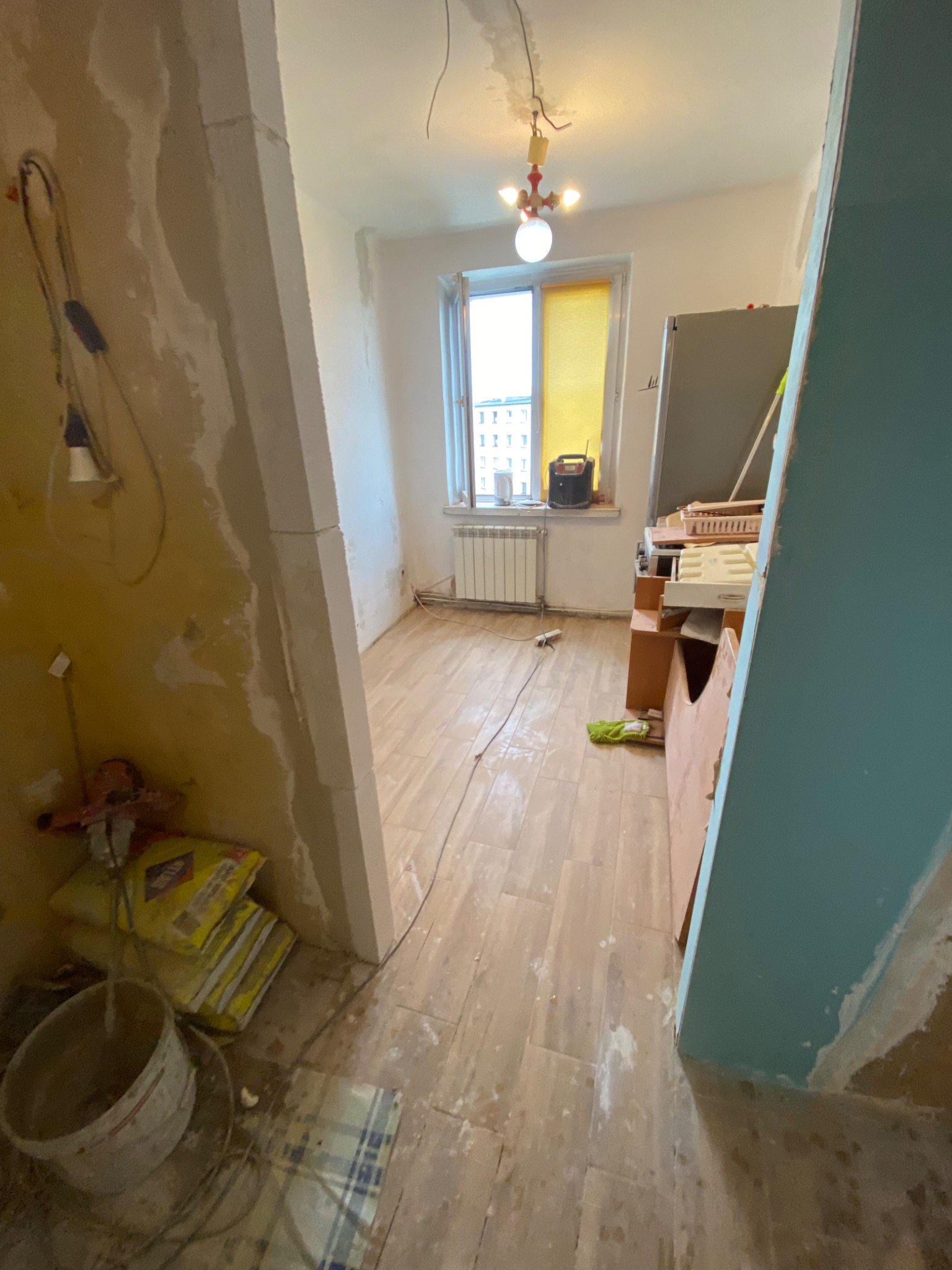 Wnętrze w trakcie remontu: widoczne worki z zaprawą, niedokończone ściany, częściowo ułożona podłoga z paneli, okno z żółtą zasłoną i grzejnik.