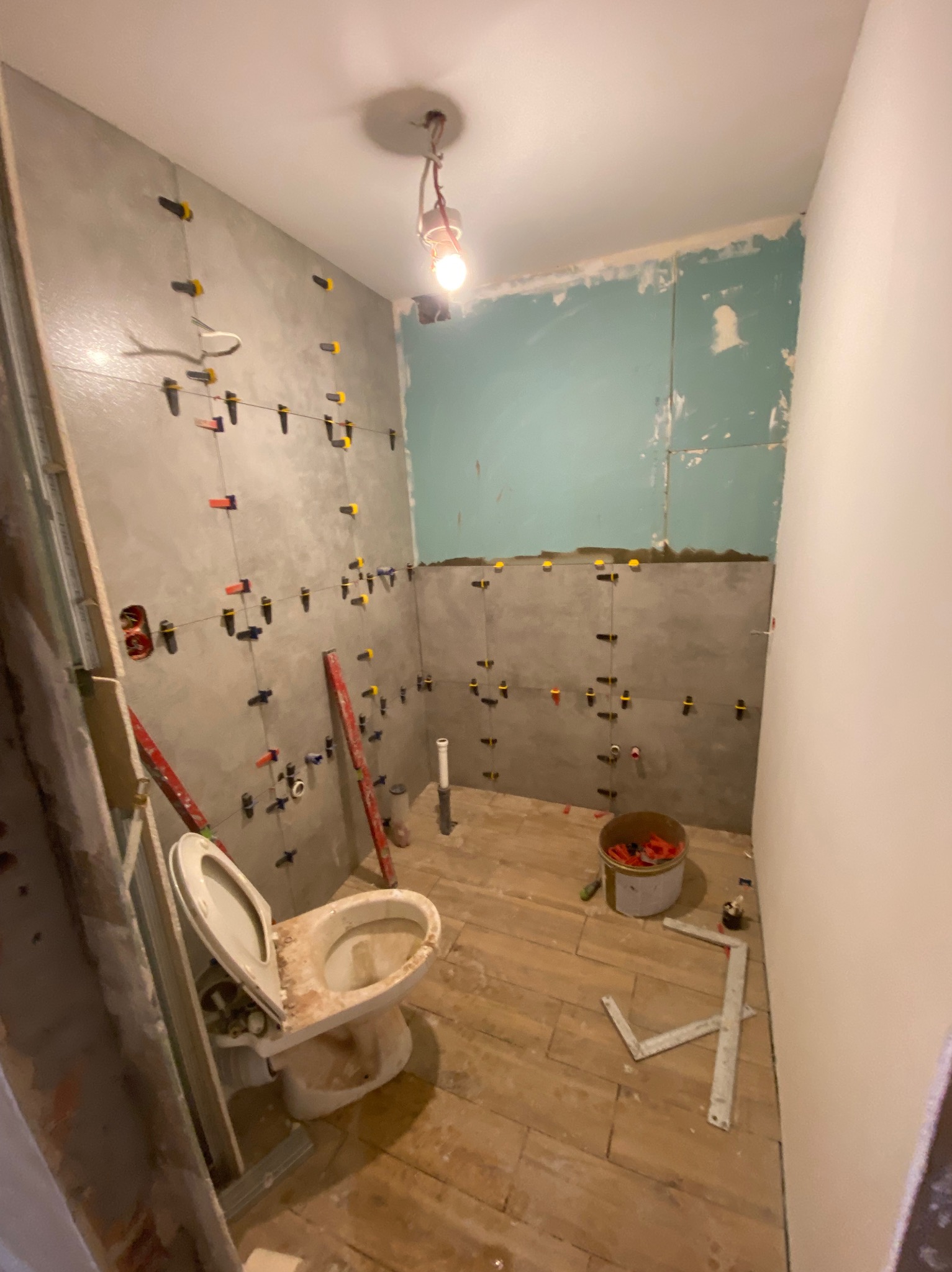 Łazienka w trakcie prac: szare płytki na ścianach z systemem poziomowania, stara toaleta, drewnopodobne płytki na podłodze. Widoczne narzędzia i materiały budowlane.