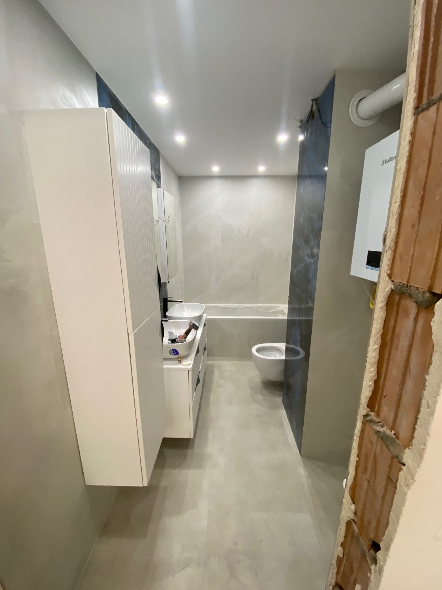 Jasna łazienka po remoncie: białe meble, umywalka, toaleta, płytki. Fragment ściany z cegły, bojler. Widok na całe pomieszczenie, neutralna kolorystyka.