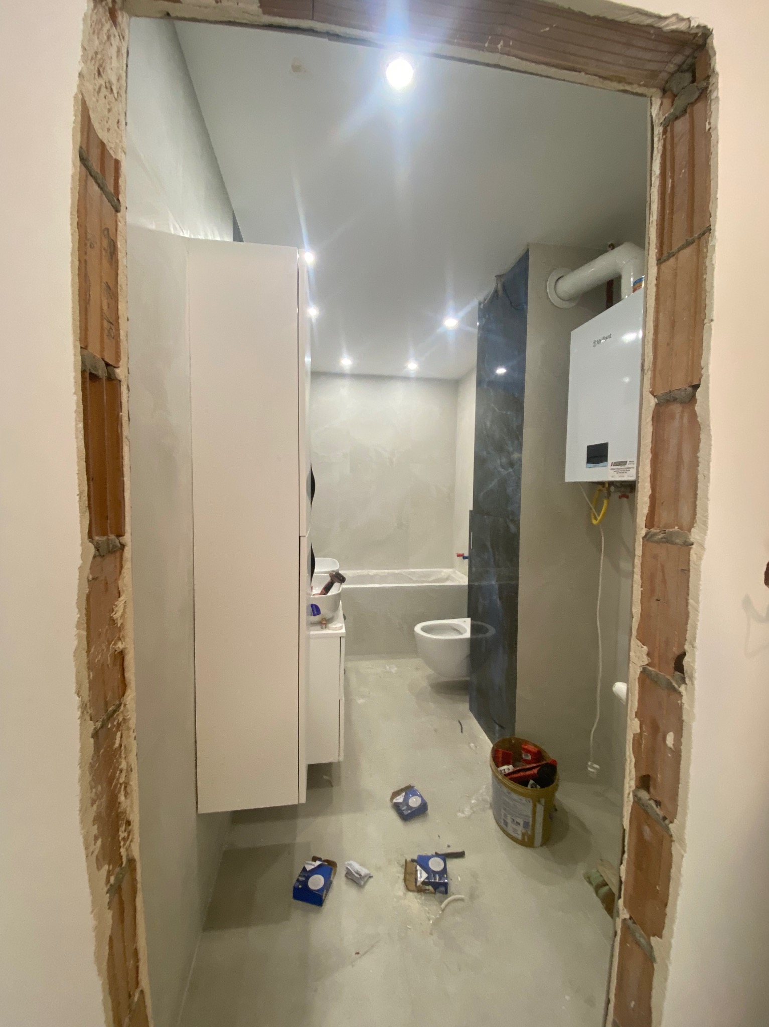 Nowoczesna łazienka w trakcie remontu, widok przez surowy otwór drzwiowy. Widoczna zabudowa, armatura, bojler i narzędzia na podłodze.