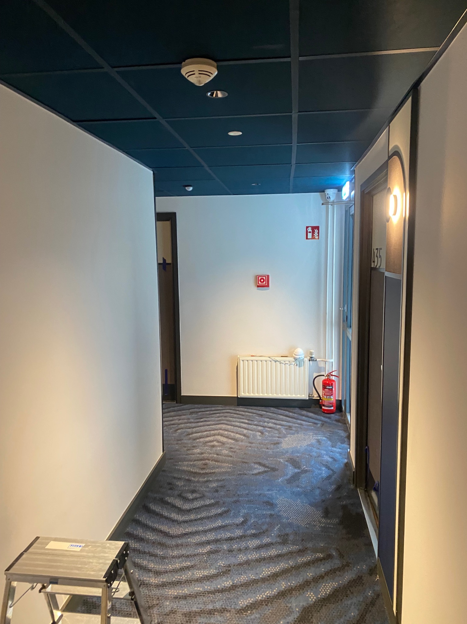 Korytarz hotelowy z wykładziną w zygzaki, grzejnikiem i gaśnicą. Widoczna drabinka malarska, sugerująca prace wykończeniowe lub konserwacyjne. Ciemna podsufitka z czujnikiem dymu.