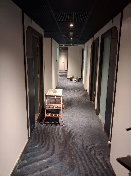 Korytarz w trakcie prac wykończeniowych: widoczny podest malarski, świeżo położona wykładzina dywanowa i pomalowane ściany. Ciemna podsufitka z kratownicą.