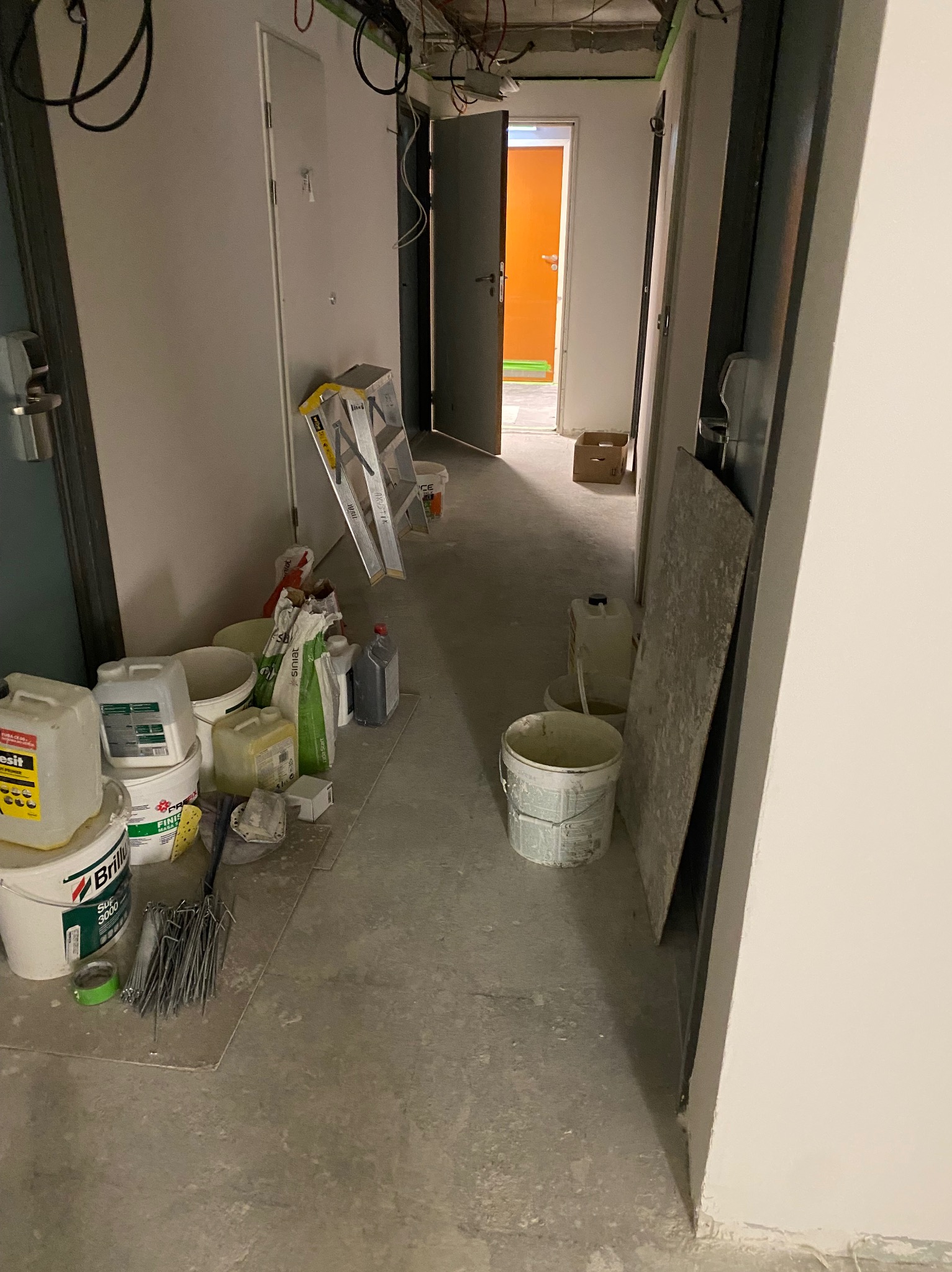 Korytarz w trakcie remontu: widoczne puszki z farbą, drabina, płyty karton-gips. Otwarta pomarańczowa ściana w tle, kable na suficie i surowa podłoga.