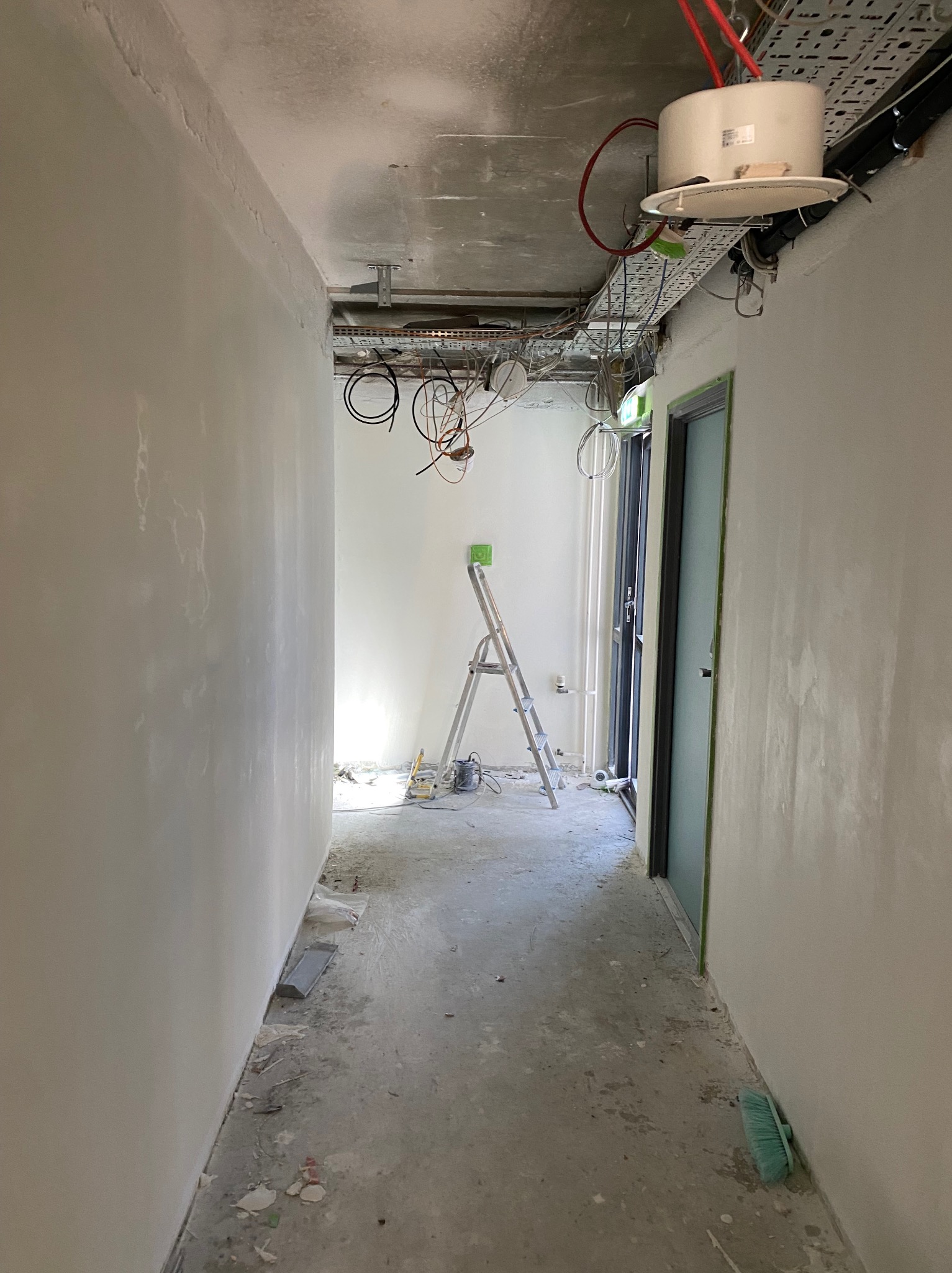 Korytarz w trakcie wygładzania ścian, widoczne ślady po gładzi, drabina i instalacje pod sufitem. Ściany przygotowane do malowania, podłoga częściowo oczyszczona.