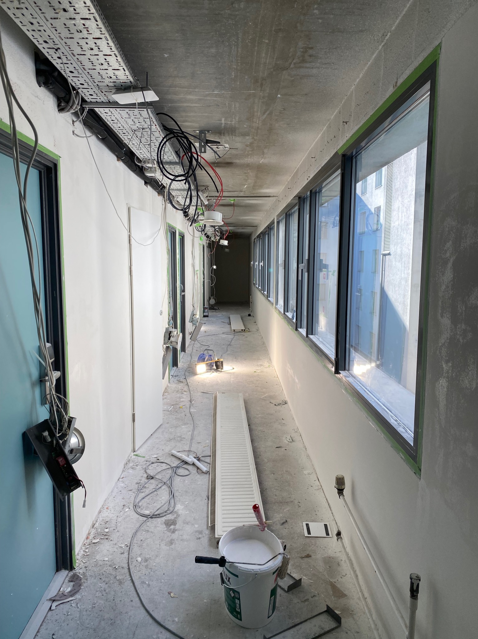 Korytarz w trakcie prac wykończeniowych: ściany przygotowane do malowania, widoczne przewody, puszki elektryczne, okna z taśmą malarską, wiadro z farbą i wałek.