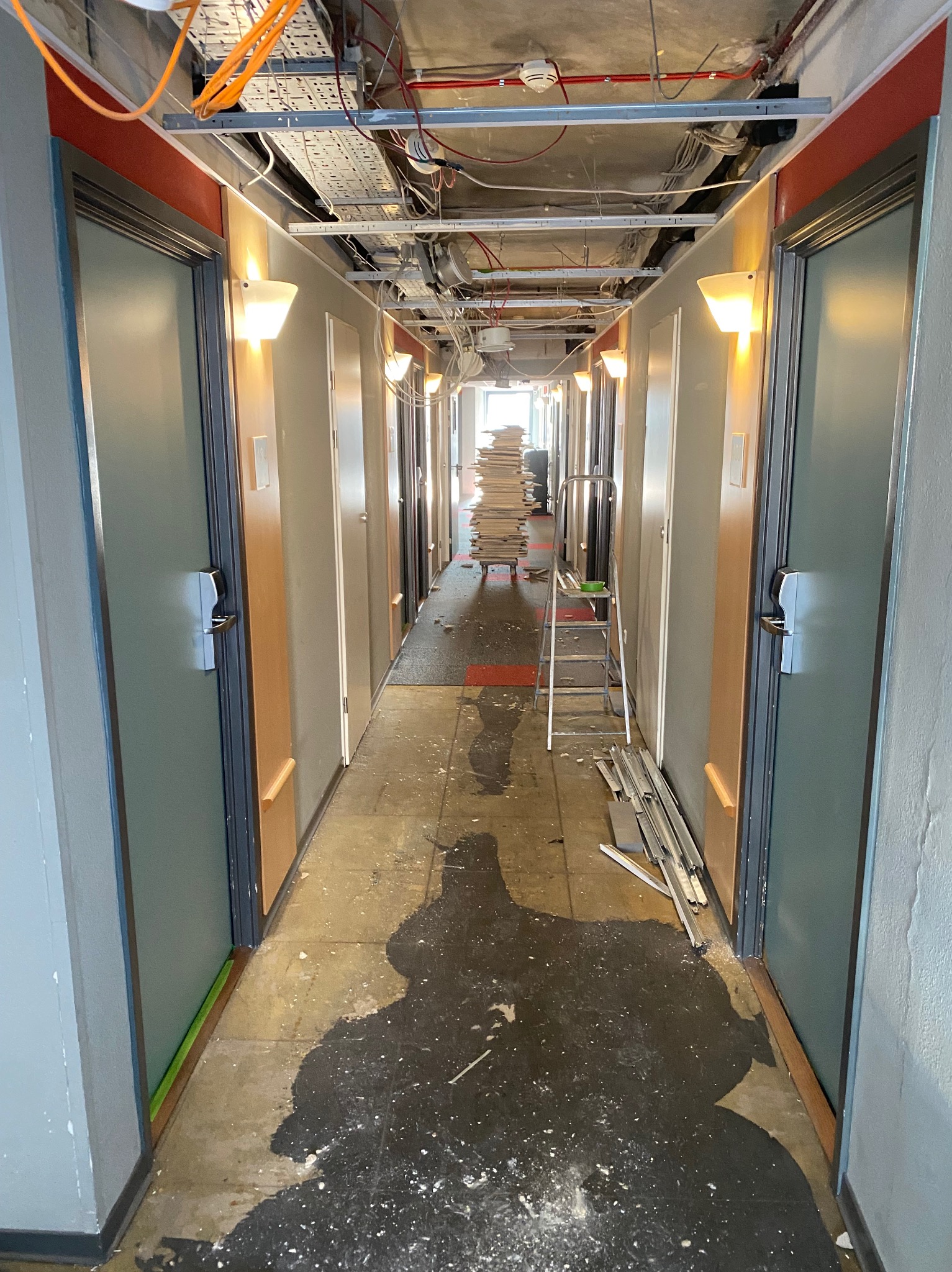 Korytarz w trakcie remontu: widoczne instalacje pod sufitem, drzwi, materiały budowlane, drabina. Na podłodze ślady po pracach wykończeniowych.