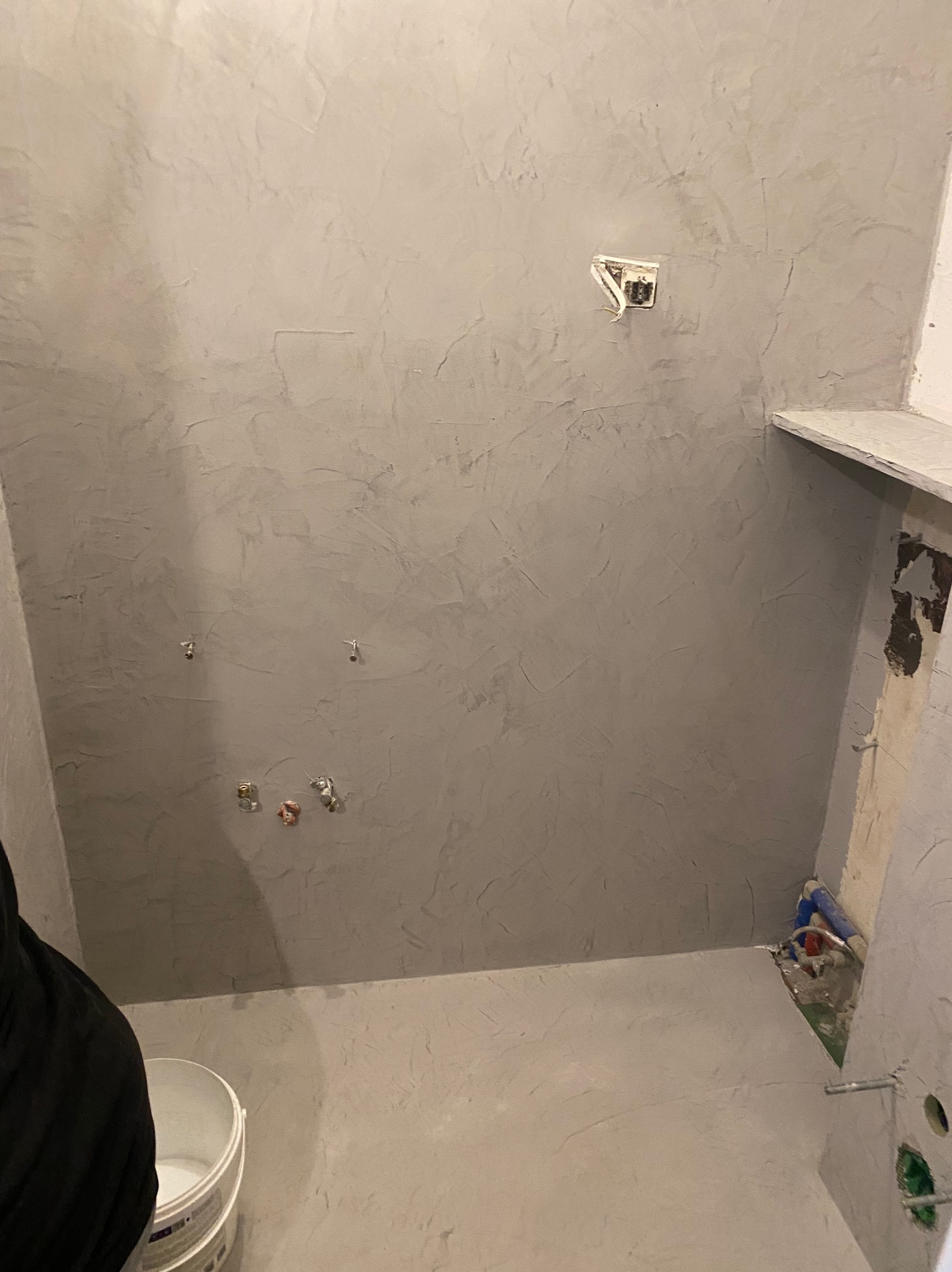 Szara, teksturowana ściana w łazience po nałożeniu gładzi, z widocznymi instalacjami i puszką elektryczną. Wiadro z farbą w rogu kadru.