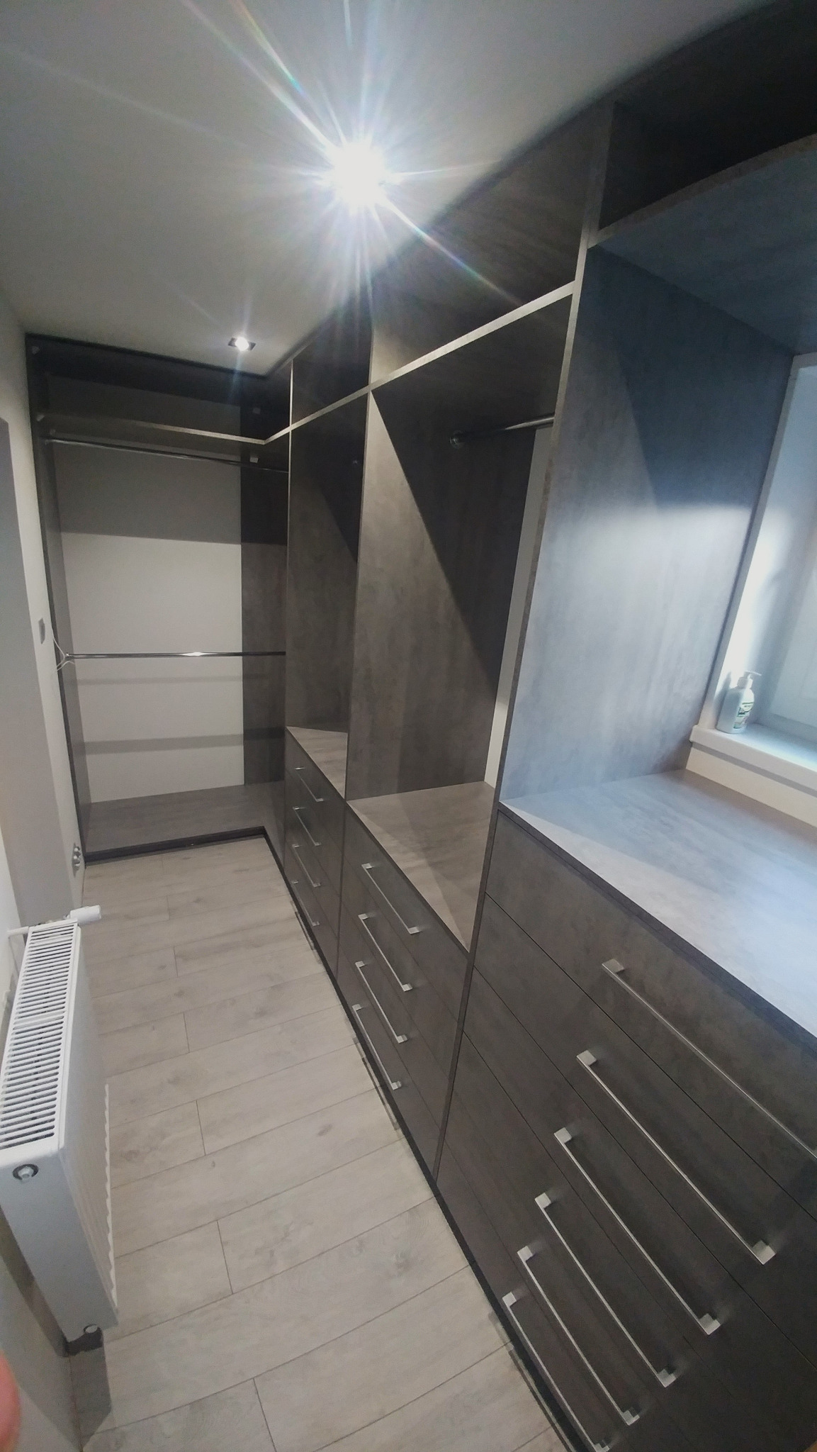 Wnętrze garderoby z szufladami i półkami w odcieniach szarości, oświetlenie punktowe w suficie, widoczny grzejnik na ścianie i drewnopodobna podłoga.