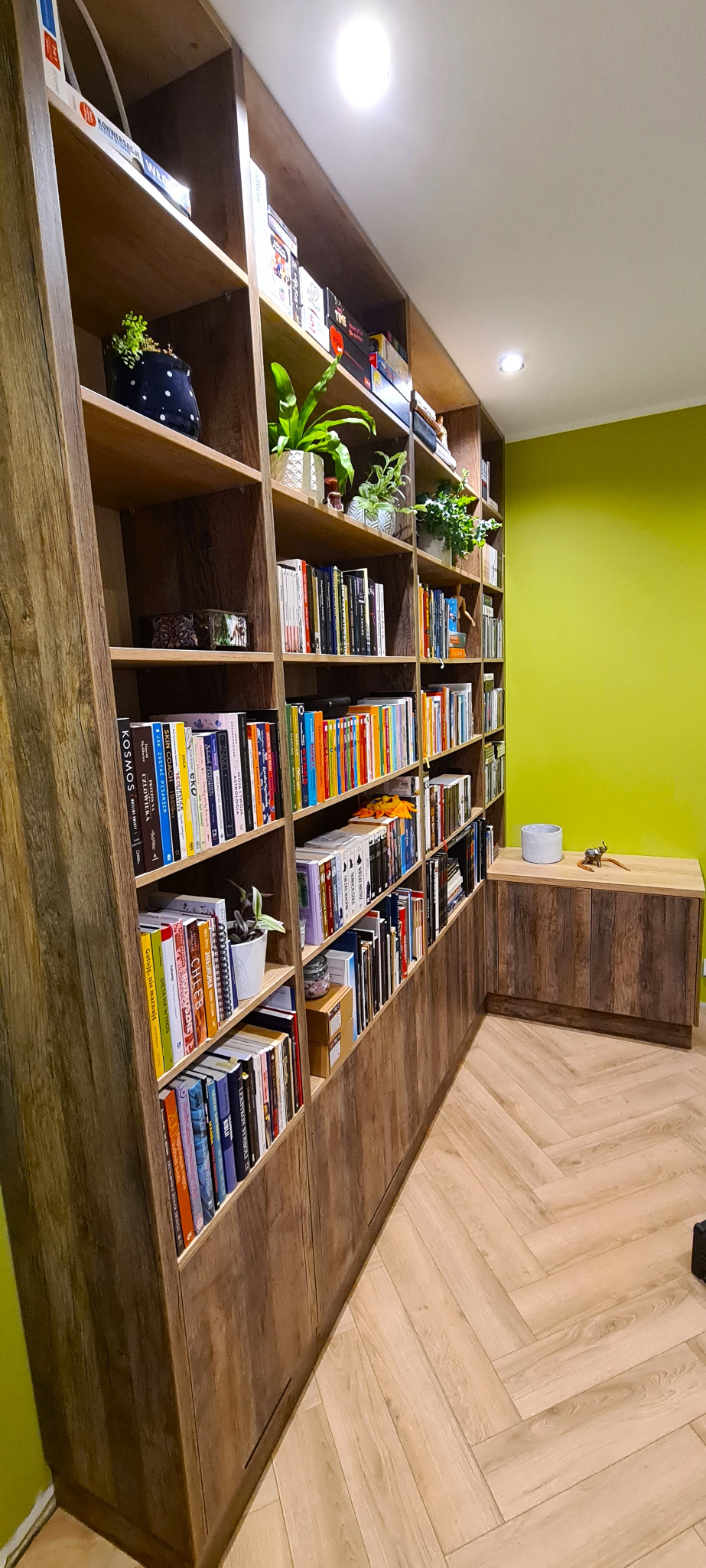 Regał na książki na wymiar z drewnianymi półkami, wypełniony książkami, roślinami doniczkowymi i dekoracjami, z szafką dopasowaną stylem i wzorem do regału, na tle zielonej ściany i parkietu...