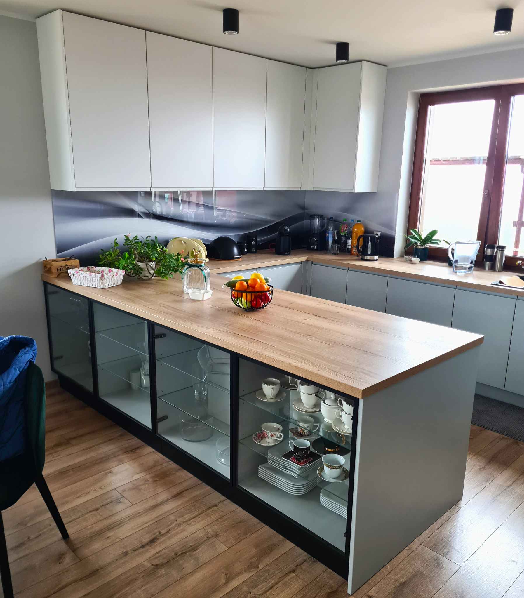 Jasna kuchnia z wyspą z przeszklonymi szafkami, na blacie o fakturze drewna owoce i dekoracje, w tle okno.