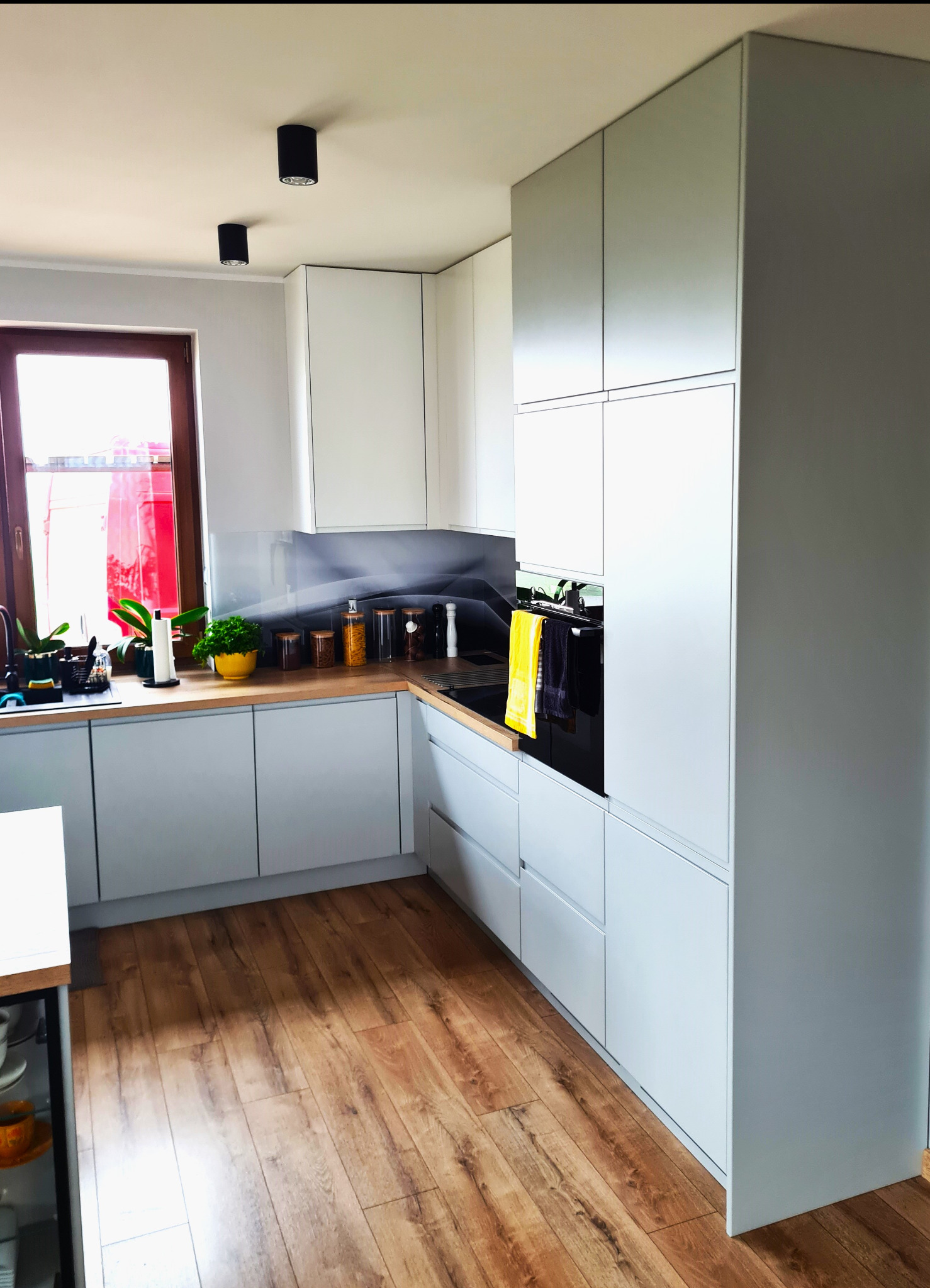 Jasnoszara zabudowa kuchenna z drewnianym blatem, okno z widokiem na czerwony samochód dostawczy, czarne lampy sufitowe