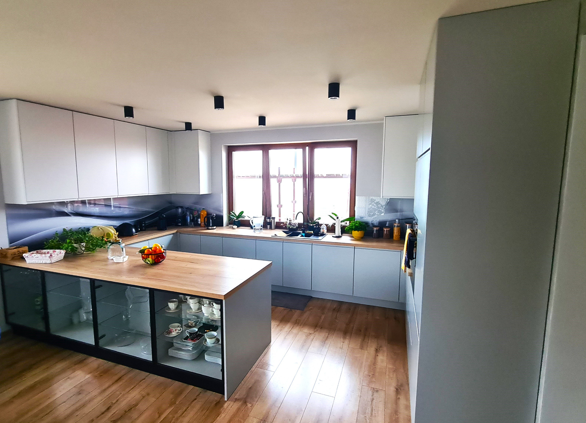 Jasna kuchnia z wyspą, drewnianym blatem i szklanymi witrynami, na blacie owoce i zioła, w tle okno.