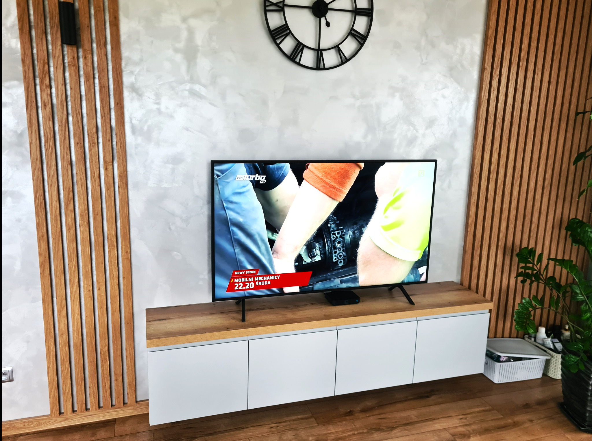 Nowoczesna szafka RTV z białymi frontami i blatem w odcieniu drewna, z telewizorem, zegarem ściennym w stylu retro i drewnianymi panelami dekoracyjnymi na ścianie w tle.