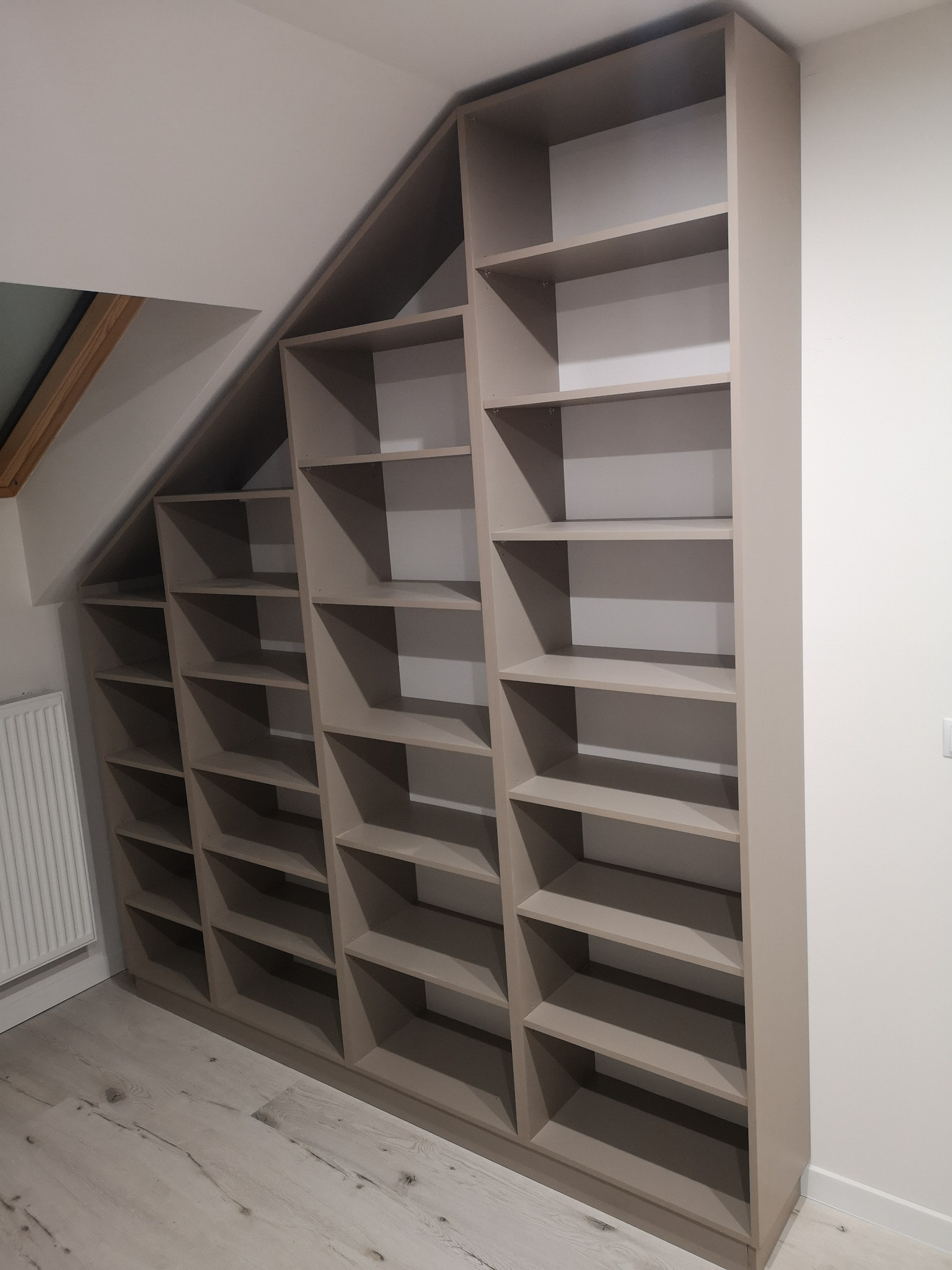 Beżowa, pusta szafa wnękowa z półkami, zabudowana pod skosem na poddaszu, z widoczną podłogą z paneli.