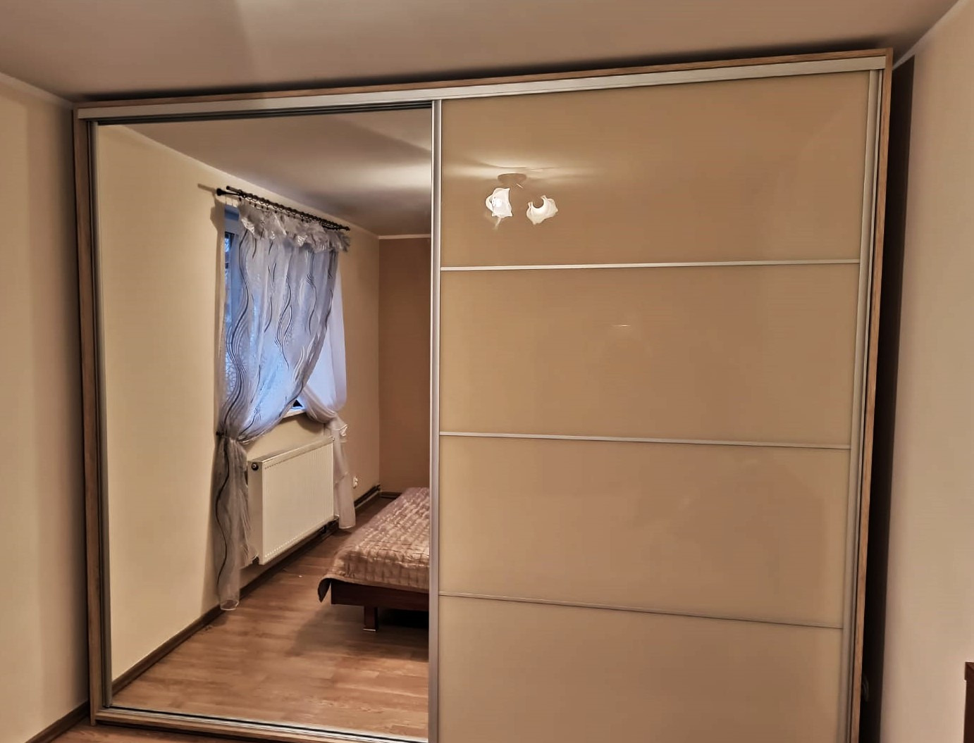 Beżowa szafa wnękowa z przesuwnymi drzwiami, jedno skrzydło lustrzane odbija sypialnię z grzejnikiem i zasłoną.