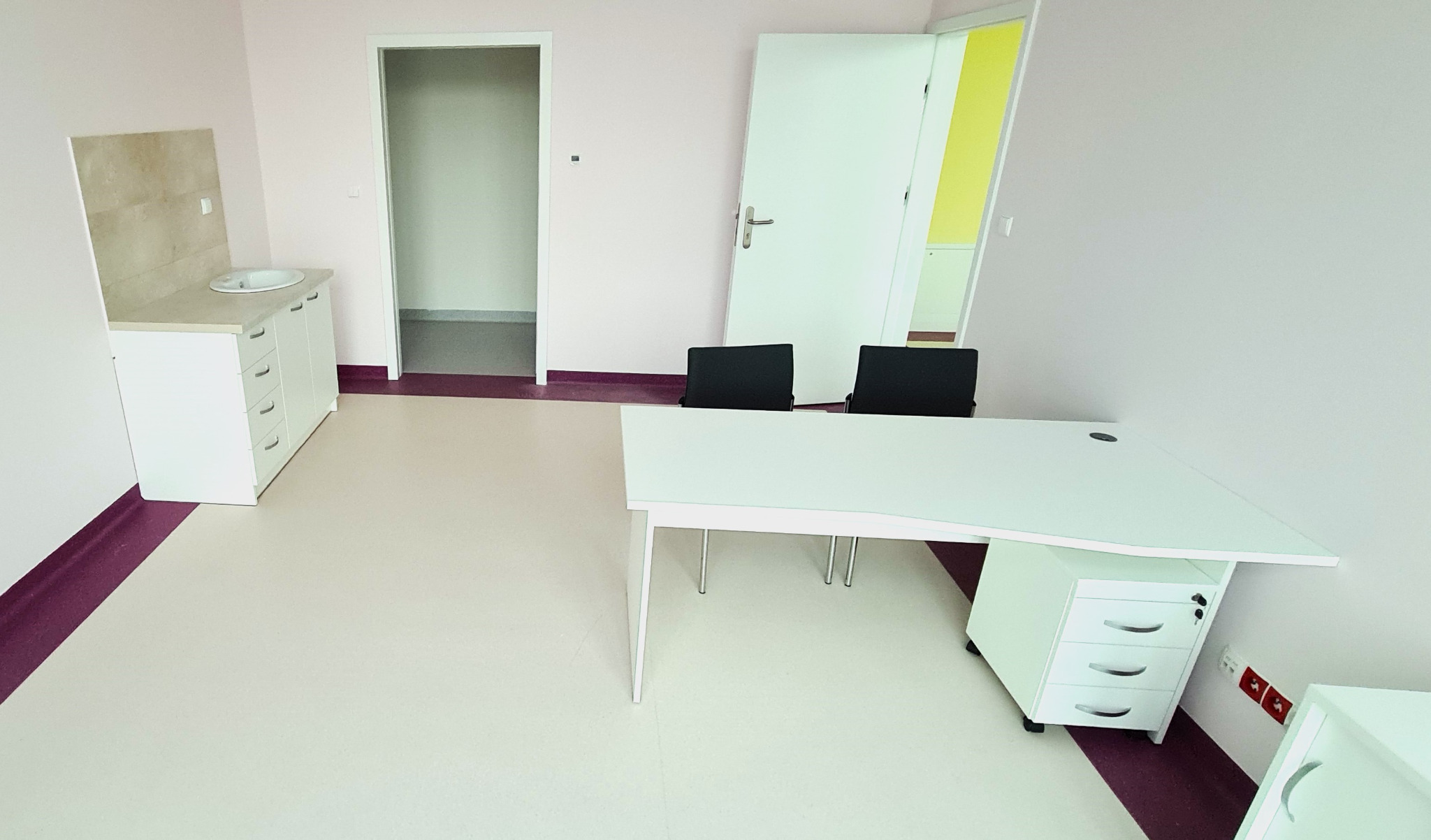 Nowoczesne wnętrze gabinetu w Kotomierzu z białymi meblami: biurko z kontenerkiem, szafka z umywalką i dwoma czarnymi krzesłami na tle jasnych ścian i podłogi z bordowymi akcentami.