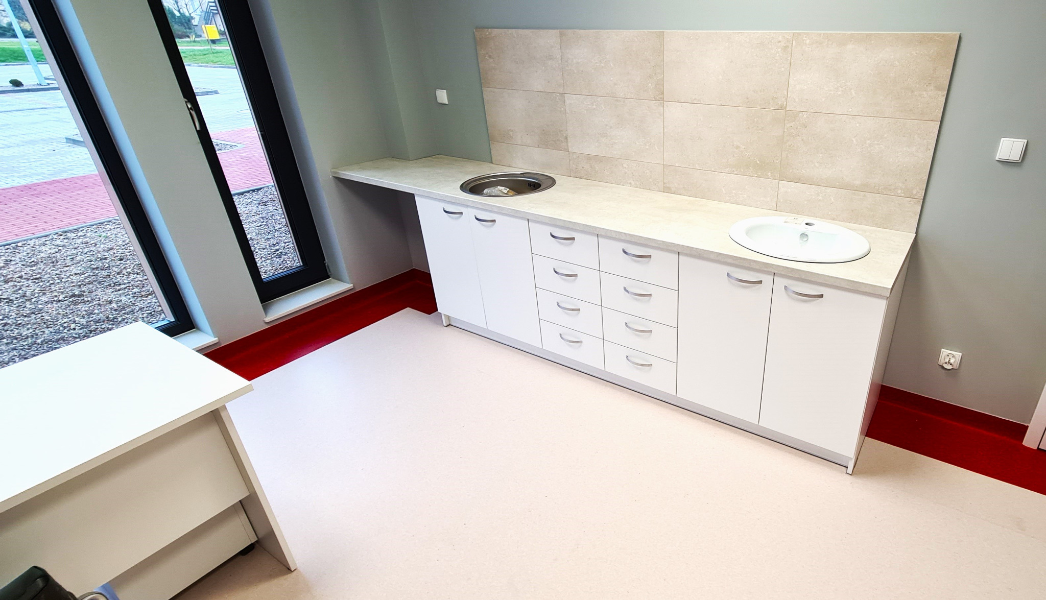 Biała zabudowa meblowa z umywalką i okrągłym zlewem, jasne płytki na ścianie, widok z góry, czerwona listwa przypodłogowa.