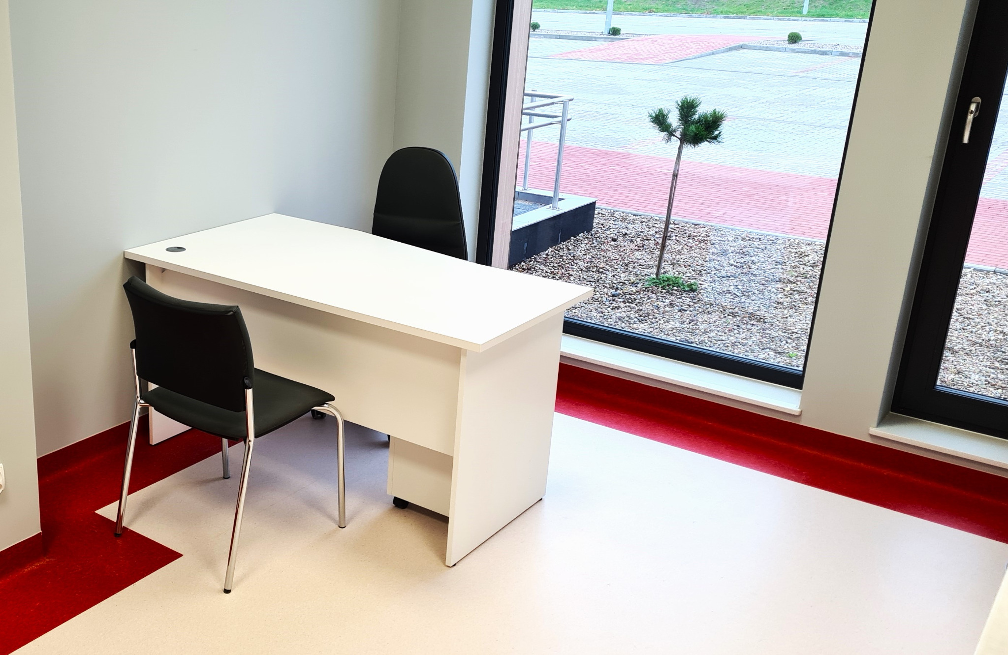 Białe biurko z czarnymi krzesłami w jasnym pomieszczeniu z dużym oknem, widok na parking i małe drzewko.