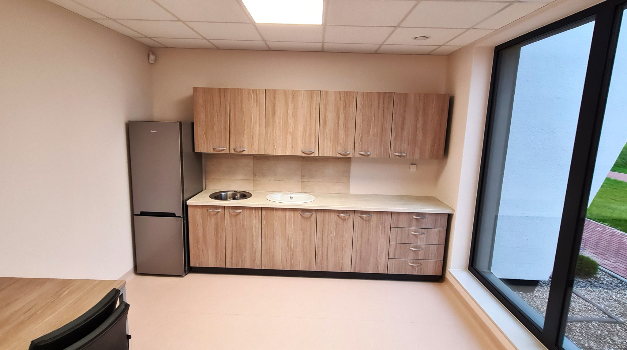 Jasna kuchnia z drewnianymi szafkami i szarą lodówką, widok z boku z dużym oknem z widokiem na zieleń.