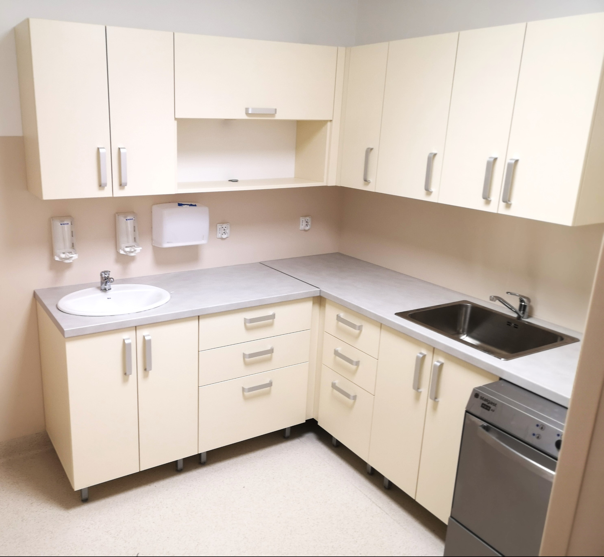 Jasna, narożna zabudowa kuchenna z beżowymi frontami i srebrnymi uchwytami, uzupełniona o zlew ze stali nierdzewnej i dozowniki na ścianie.