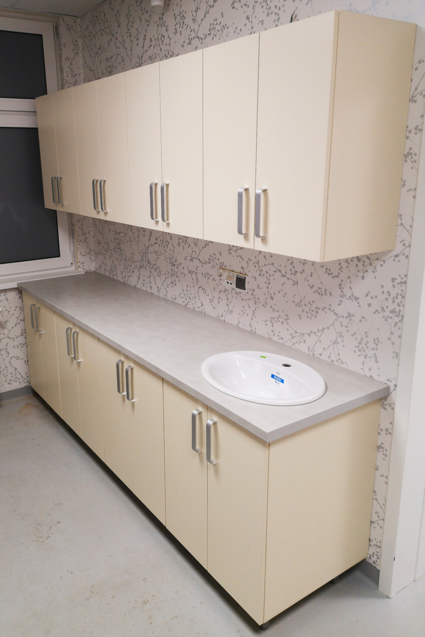 Beżowe szafki kuchenne na wymiar z szarym blatem i białą umywalką, srebrne uchwyty, tło stanowi ściana z motywem roślinnym.