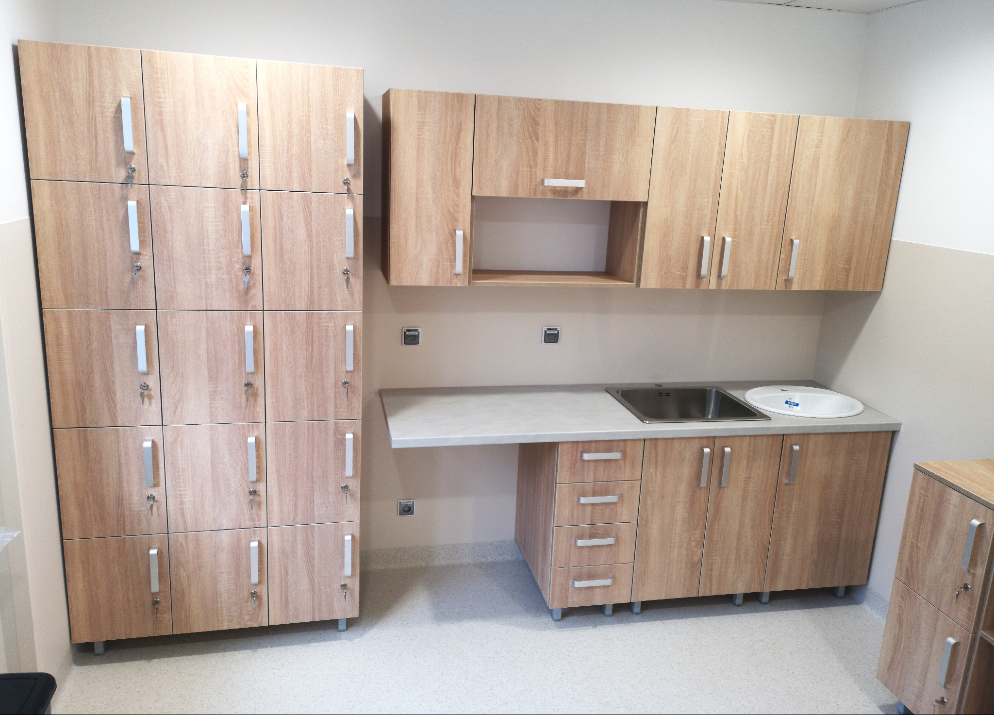 Jasne, drewnopodobne szafki z umywalką i szafa zamykana na klucz, prawdopodobnie w przestrzeni publicznej.