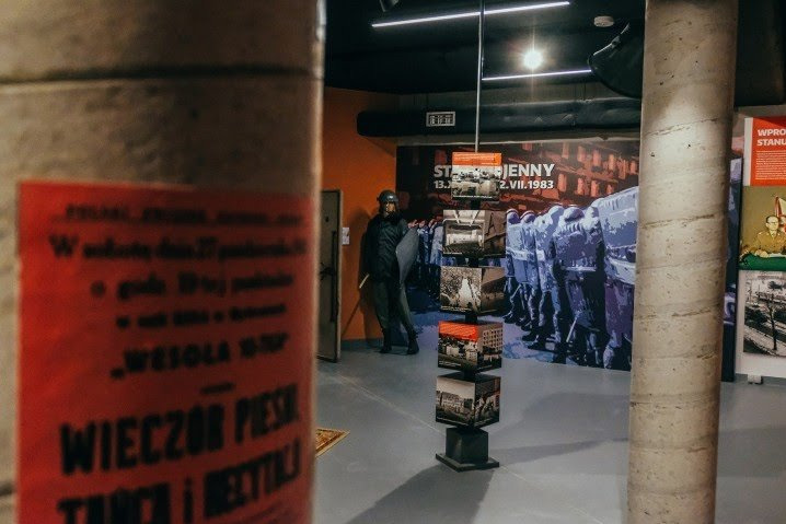 Ekspozycja muzealna: plakat z napisem 'Wieczór pieśni, tańca i dechty' na tle instalacji przedstawiającej oddział ZOMO w czasie stanu wojennego, z figurą funkcjonariusza w hełmie i z tarczą.