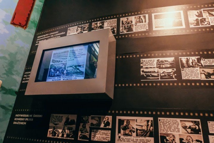 Ekspozycja muzealna prezentująca informacje o obronie cywilnej, z ekranem wyświetlającym archiwalne zdjęcia lotniczych bomb zapalających i lotniczych pozrzutów wylewczych ITP, otoczonym grafikami...