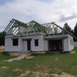 Surowy stan domu z białego bloczka, widoczne otwory okienne i drzwiowe, zielona konstrukcja dachu w trakcie montażu, teren wokół zarośnięty trawą.