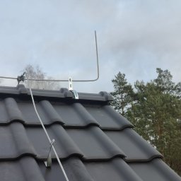 Easy Elektrik Mateusz Iwanowicz - Ujęcie z dołu na fragment dachu z czarnej dachówki ceramicznej, na którym zamontowano metalowy maszt odgromowy z przewodem, uchwyconym metalowymi klamrami. W tle widoczne drzewa i pochmurne niebo.