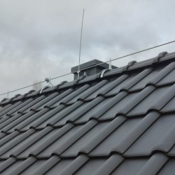 Easy Elektrik Mateusz Iwanowicz - Szare dachówki ceramiczne z zamontowaną instalacją odgromową: przewód uziemiający z uchwytami i maszt odgromowy na tle pochmurnego nieba.