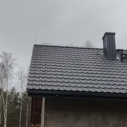 Easy Elektrik Mateusz Iwanowicz - Szary dach z metalowej dachówki z zamontowaną instalacją odgromową, widoczny komin i fragment ściany budynku w trakcie wykończenia, tło stanowią drzewa i pochmurne niebo.