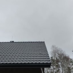 Easy Elektrik Mateusz Iwanowicz - Szary dach z dachówki ceramicznej z zamontowaną instalacją odgromową, widoczne przewody i maszty, w tle drzewa i pochmurne niebo.