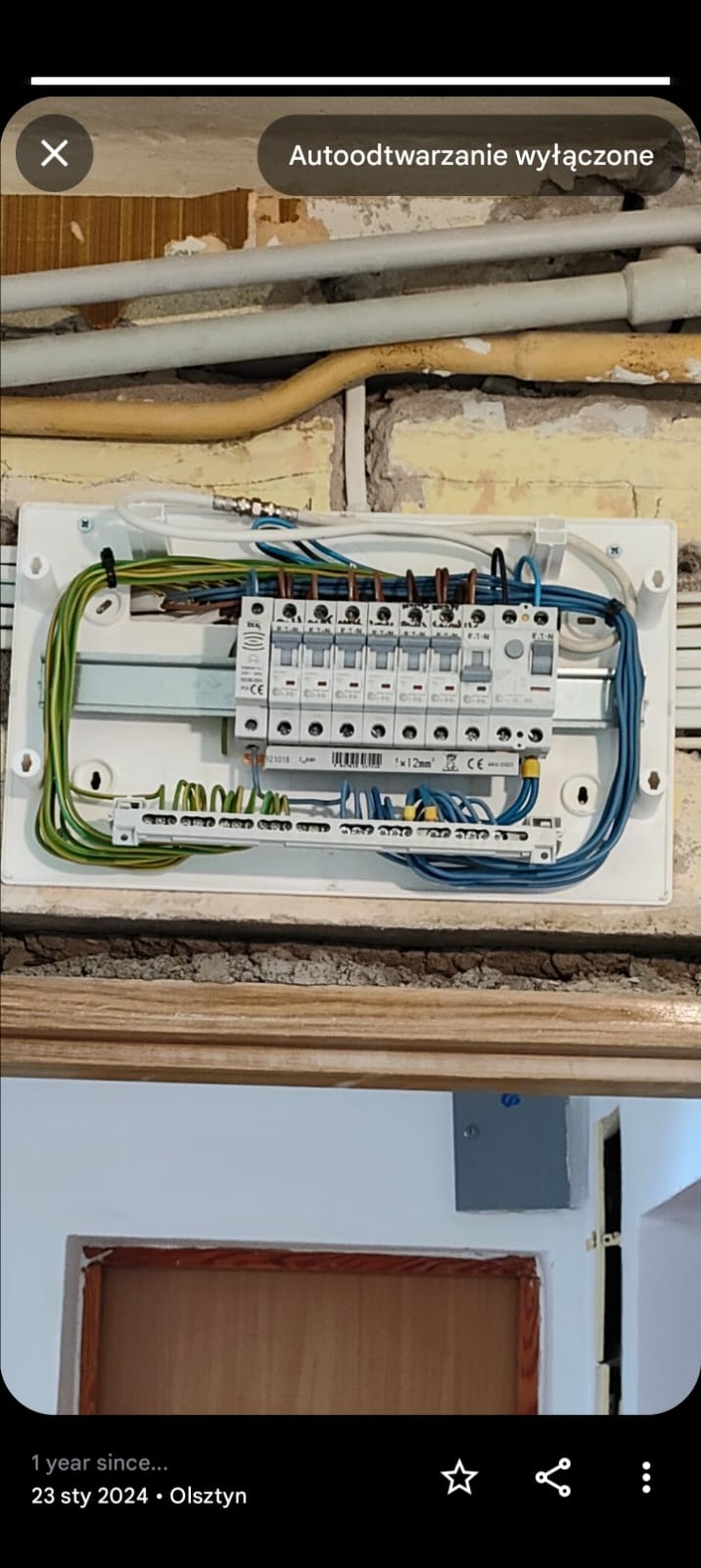 Rozdzielnica elektryczna z zabezpieczeniami, widoczne przewody w kolorach zielono-żółtym, niebieskim i brązowym, zamontowana na ścianie z widocznymi przewodami i rurami instalacyjnymi, częściowo...