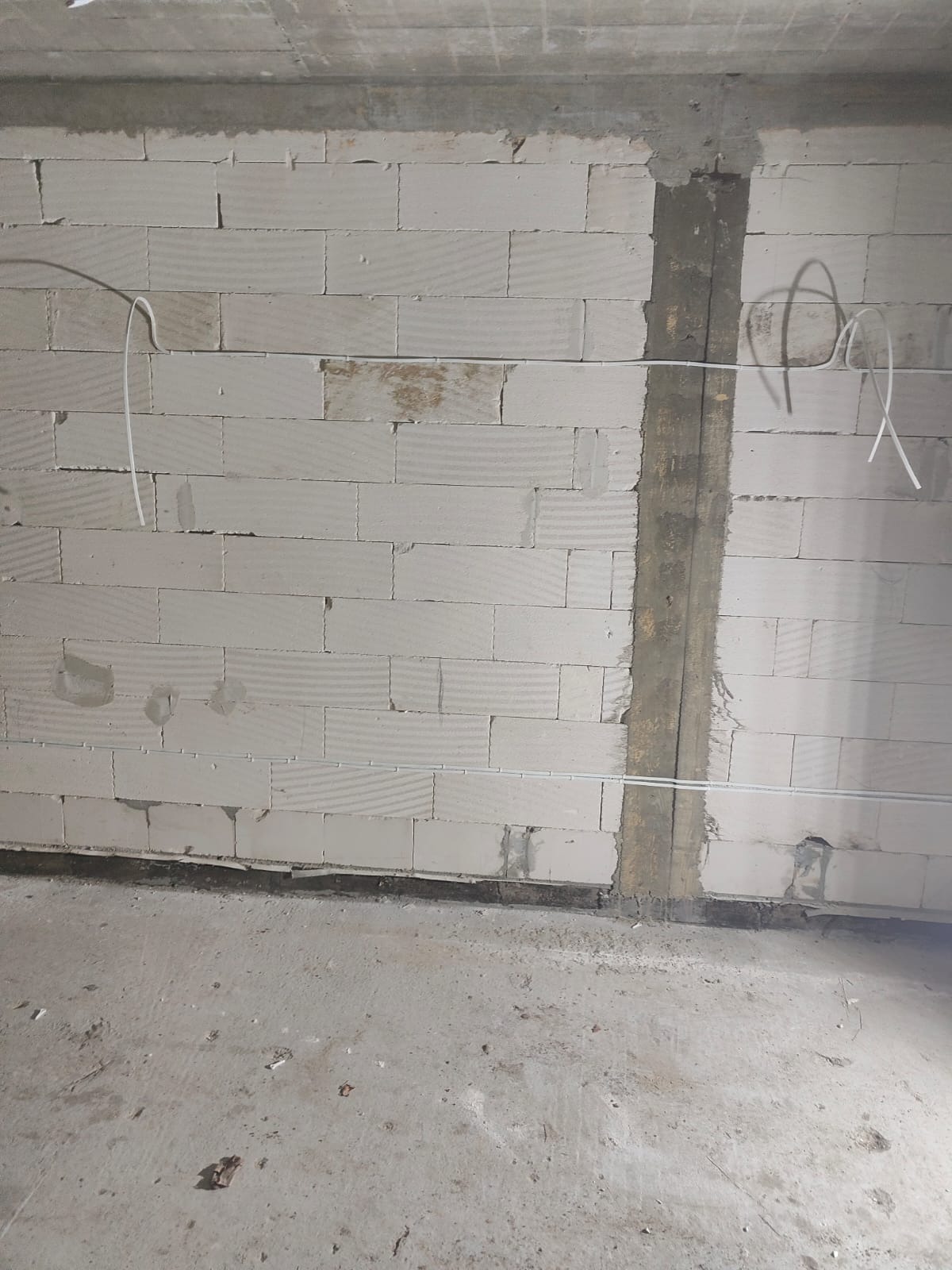Ściana z betonu komórkowego z pionowym bruzdą wypełnioną zaprawą, przez którą poprowadzone są kable elektryczne w białych peszlach, z widocznymi fragmentami kabli wystającymi ze ściany.